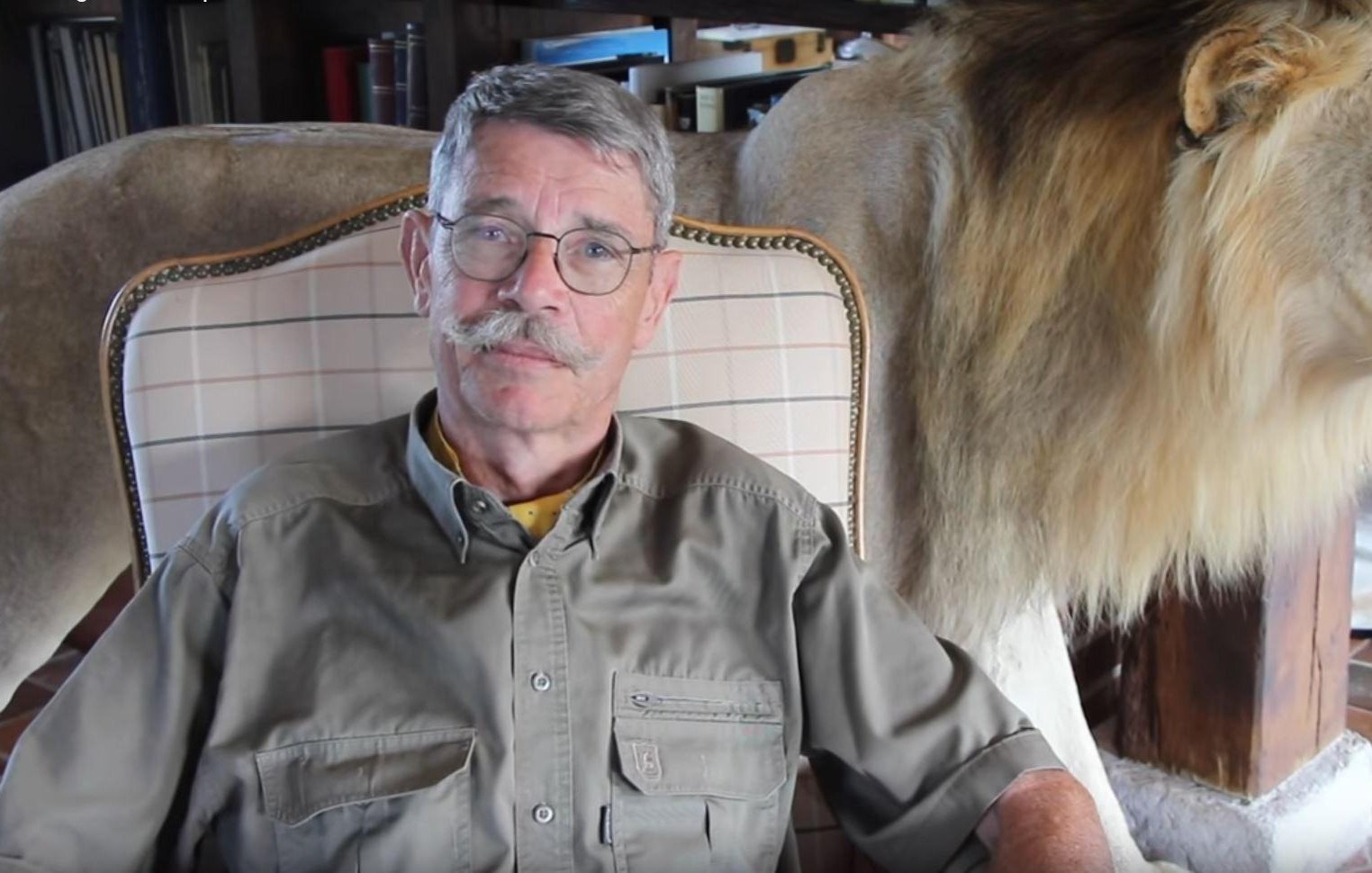 L'image montre un homme assis sur un fauteuil. Il a des lunettes, une moustache grise et porte une chemise kaki. À côté de lui, il y a un lion naturalisé, avec une crinière fournie et un regard majestueux. Le cadre donne une impression chaleureuse et rustique, avec une ambiance intérieure qui suggère une maison ou un chalet. Le contraste entre l'homme et le lion évoque une connexion avec la nature sauvage.