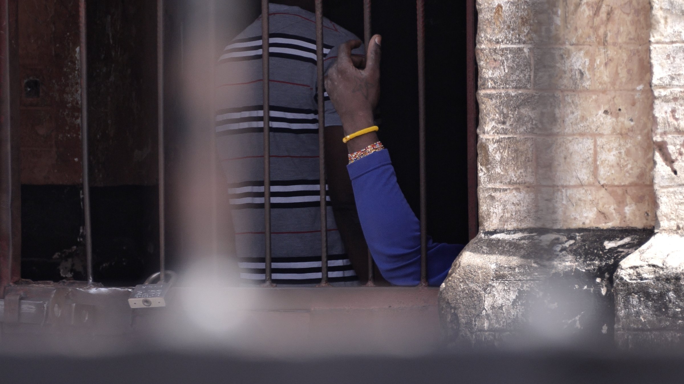 Cette image montre une scène à l'intérieur d'un espace confiné, avec des barreaux visibles au premier plan. On distingue des silhouettes humaines derrière les barreaux. Une main, portant un bracelet, s'élève légèrement, témoignant d'un geste de communication ou de contact. Les murs autour des barreaux semblent anciens et en pierre, ajoutant un élément d'authenticité à l'environnement. L'éclairage suggère que la scène se déroule à l'extérieur, avec une lumière naturelle qui éclaire partiellement l'espace.