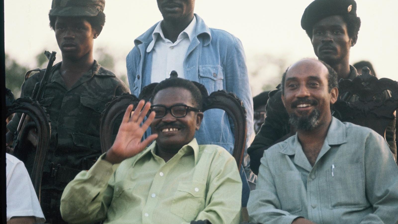 L'image présente un groupe de personnes, dont deux hommes au premier plan, assis sur des chaises ornées. L'un des hommes, souriant et portant des lunettes, salue de la main. Il a une chemise verte claire. À sa droite, un autre homme avec une chemise beige sourit également. En arrière-plan, plusieurs soldats en uniforme de camouflage et avec des casquettes se tiennent debout. L'environnement semble être en plein air, peut-être lors d'un événement officiel, avec un ciel illuminé par le crépuscule.