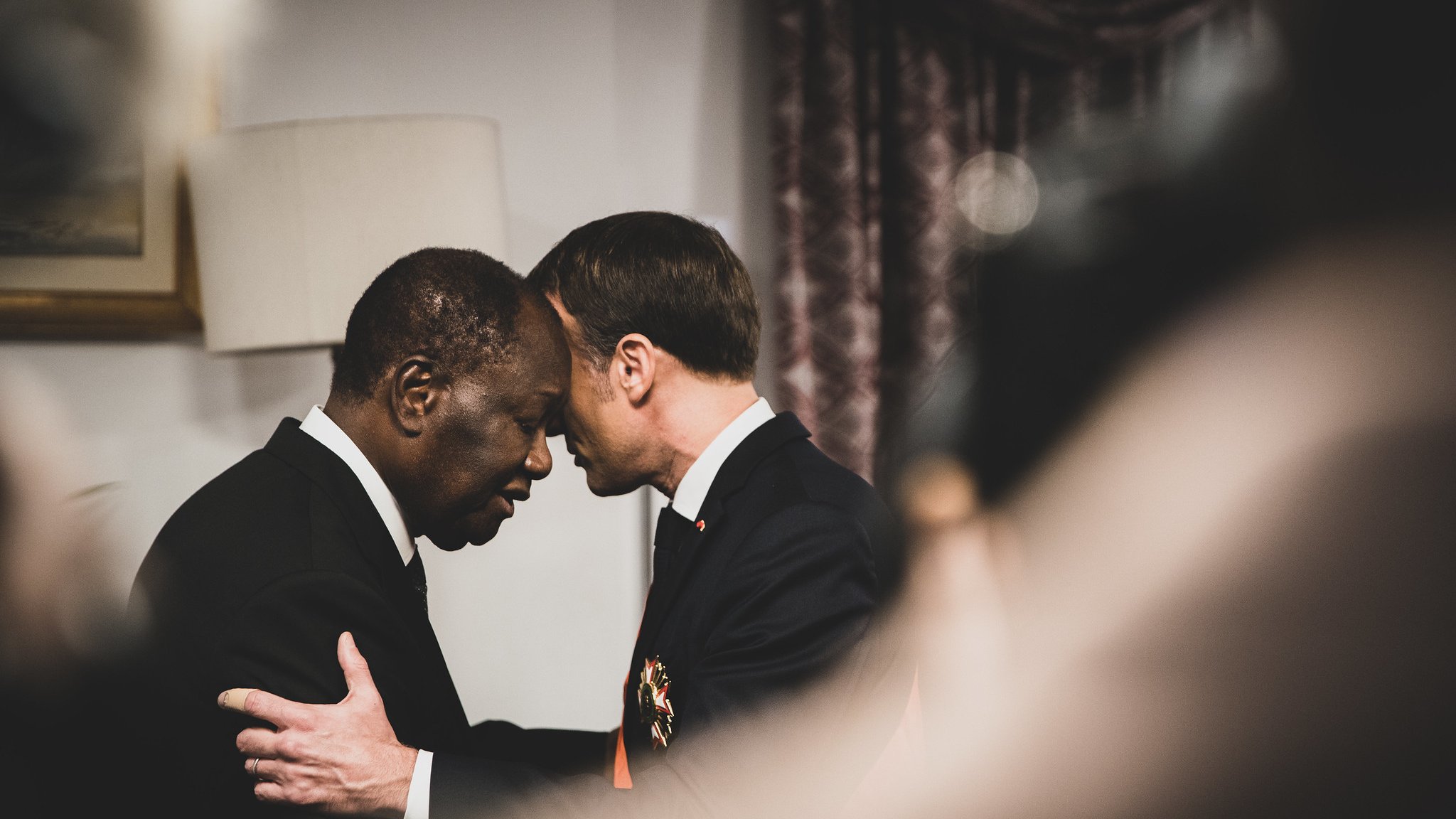 L'image montre deux hommes en train de se saluer de manière chaleureuse, leurs fronts se touchant, ce qui suggère une connexion profonde ou un moment d'émotion. L'un des hommes porte un costume sombre, tandis que l'autre semble également en habit formel. Le décor en arrière-plan, légèrement flou, indique un environnement intérieur, probablement un bureau ou une salle de conférences. La lumière est douce et crée une atmosphère respectueuse et intime. Les expressions des deux hommes semblent sérieuses et significatives, évoquant un échange important ou une reconnaissance mutuelle.