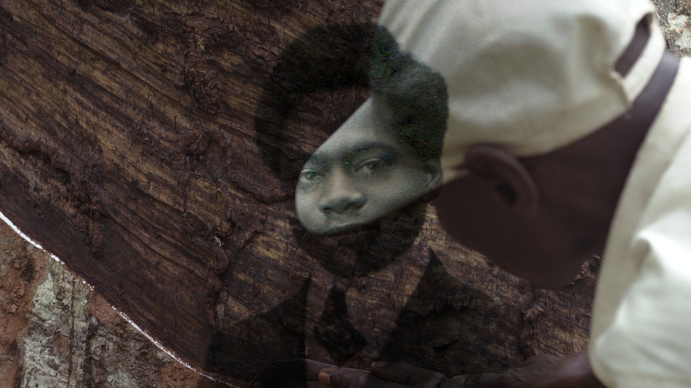 L'image montre un homme en train de travailler avec un morceau de bois. Ce dernier est une surface très texturée, et l'homme semble utiliser un outil pour façonner ou sculpter le bois. À travers le bois, un portrait en noir et blanc de quelqu'un d'autre est visible, créant un effet de superposition. Le visage qui apparaît dans le bois a une expression sérieuse, et l'homme qui travaille porte une casquette. L'atmosphère de l'image évoque une connexion entre le passé et le présent, comme si l'artisan essayait de donner vie à la mémoire de cette personne.