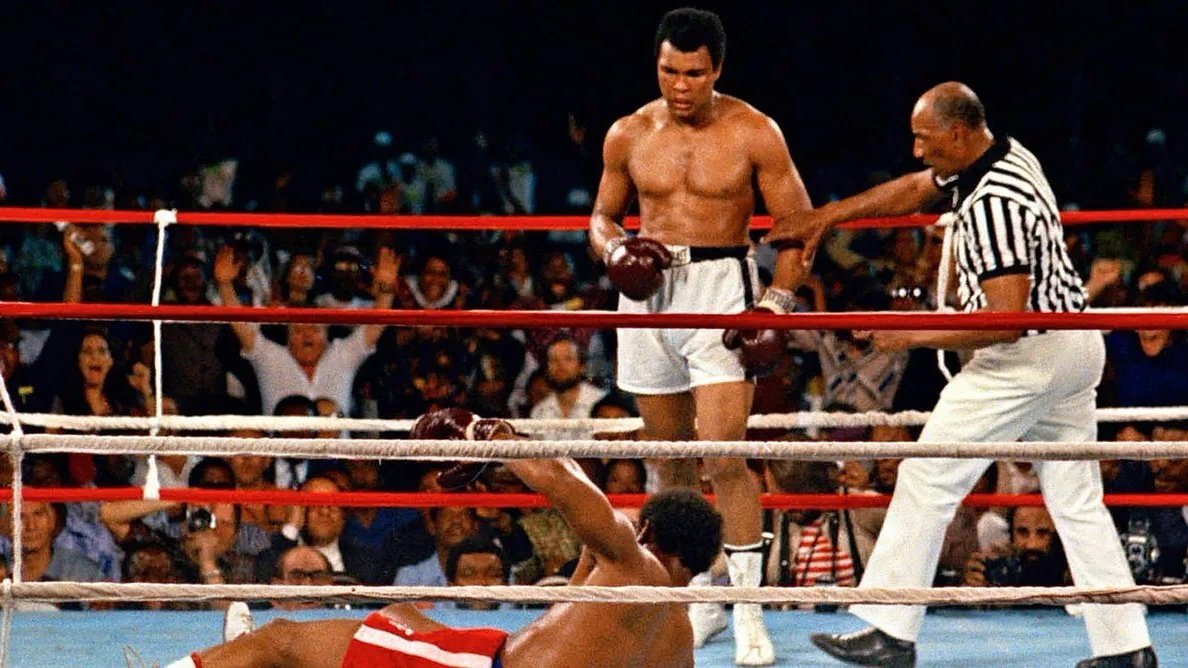 L'image montre un moment mémorable d'un match de boxe. Au centre, un boxeur, torse nu et portant des shorts blancs, se tient debout avec un regard déterminé. C'est Muhammad Ali, connu pour sa rapidité et sa technique. À ses pieds, un autre boxeur est allongé sur le sol, faisant face au ring, après avoir été mis au tapis. Le public en arrière-plan est en émoi, certains levant les bras en signe d'enthousiasme ou d'étonnement. Un arbitre, vêtu d'un maillot à rayures, s'approche du boxeur au sol, prêt à commencer le compte. L'atmosphère est chargée d'intensité et d'adrénaline, typique des combats de boxe.