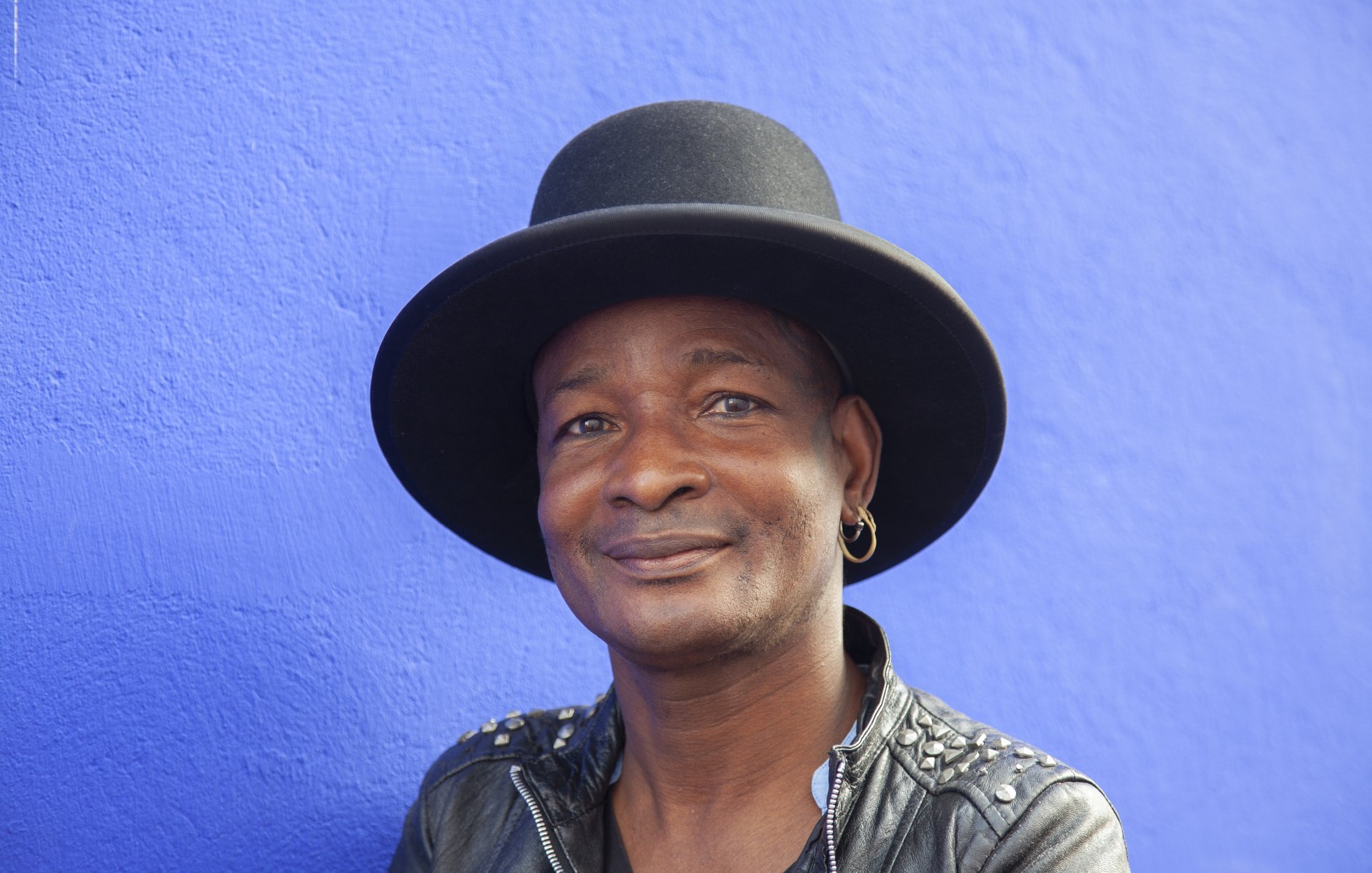 Cette image montre un homme souriant, portant un grand chapeau noir qui a une forme de haut de forme. Il est habillé d'une veste en cuir noire ornée de clous, ce qui lui donne un style à la fois décontracté et audacieux. L'arrière-plan est d'un bleu vif, créant un contraste frappant avec ses traits. Son expression est amicale et accueillante, évoquant une personnalité chaleureuse et charismatique.