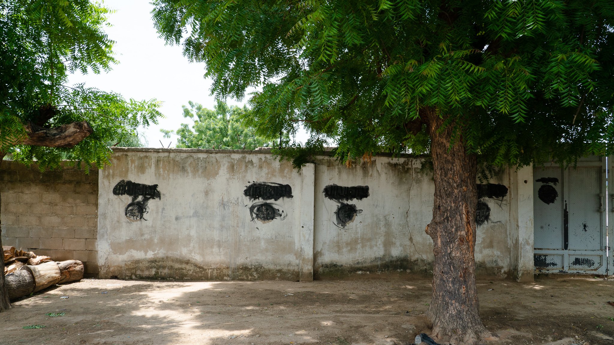 L'image présente un mur en béton aux couleurs neutres, probablement gris ou beige, partiellement recouvert par des ombres de feuillage provenant d'un grand arbre à proximité. Sur le mur, on peut voir des traces de peinture noire qui forment des motifs stylisés évoquant des yeux. Ces motifs sont positionnés de manière à créer une disposition particulière sur le mur. Au premier plan, on remarque également un tronc d'arbre robuste, avec des racines visibles qui émergent du sol sablonneux. L'environnement semble tranquille, avec une légère luminosité qui indique une journée ensoleillée.
