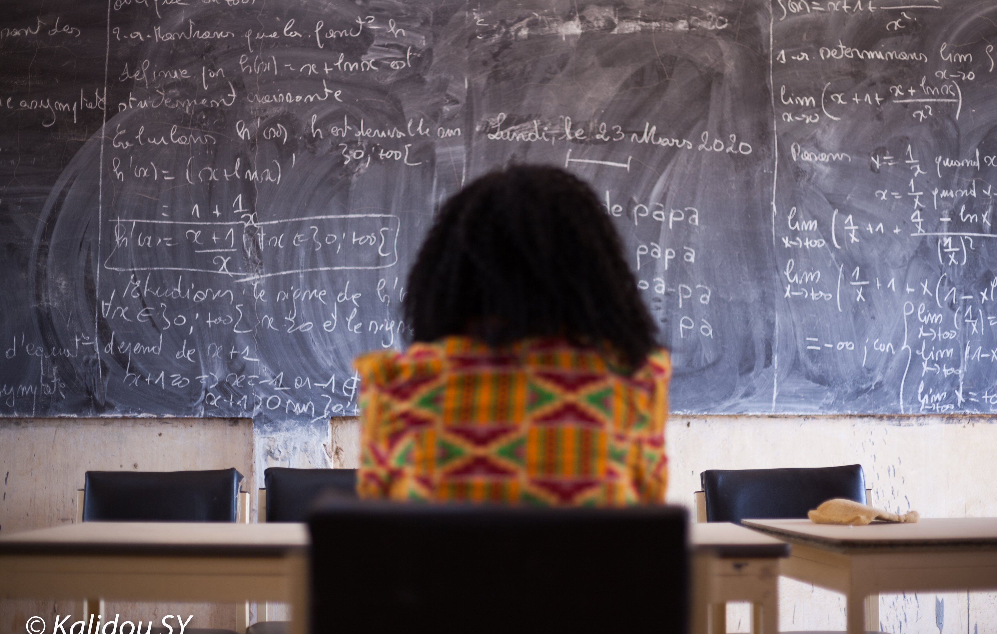 Dans cette image, on voit une personne assise, de dos, face à un grand tableau noir couvert d'écritures en craie blanche. Les formules mathématiques et les notes semblent complexes, suggérant un environnement académique ou d'apprentissage. La personne porte un vêtement coloré avec des motifs vibrants, ajoutant une touche de culture. L'ambiance est studieuse, et l'environnement évoque une salle de classe ou un espace de travail. Le contraste entre les couleurs vives du vêtement et la sombre surface du tableau crée une composition visuelle intéressante.