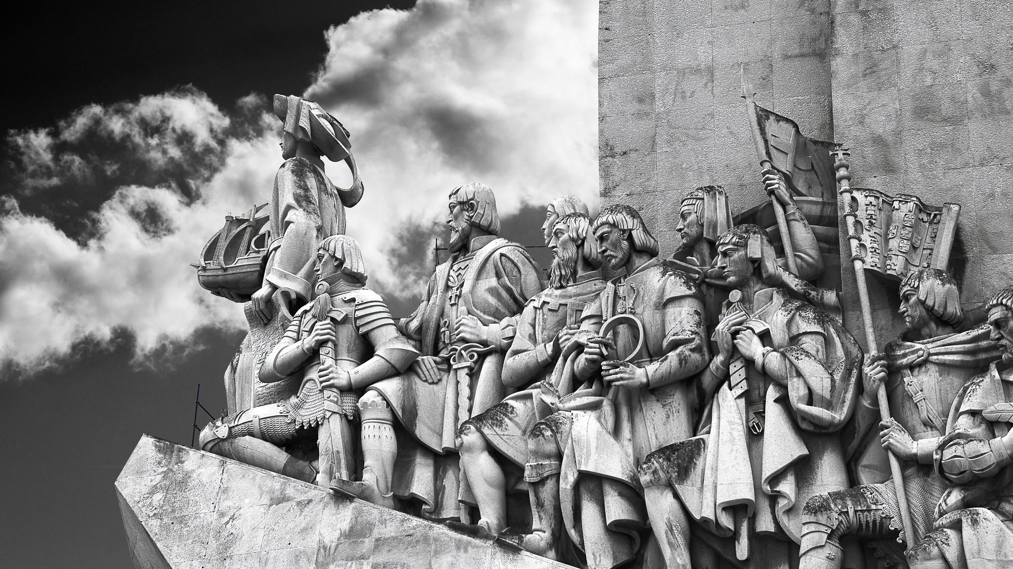 L'image représente un relief sculpté monumentale. On y voit une procession de figures humaines, probablement des explorateurs ou des conquérants, qui se tiennent debout sur une plateforme. Les personnages sont vêtus de costumes historiques et affichent des expressions déterminées. Certains d'entre eux portent des armes, des drapeaux et d'autres objets symboliques, témoignant de leurs exploits. En arrière-plan, on aperçoit des nuages sombres qui ajoutent une atmosphère dramatique à la scène. Le tout est sculpté dans un matériau clair, probablement de la pierre, permettant de bien contraster avec le ciel nuageux. Cette œuvre dégage une impression de puissance et de grandeur, évoquant une célébration de l'histoire et de l'aventure.