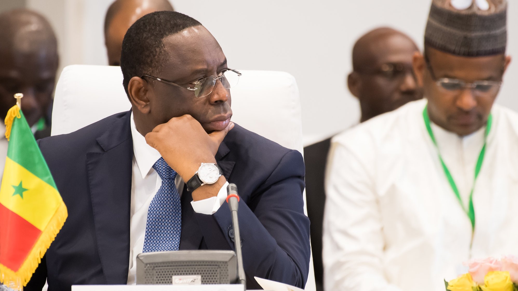 L'image montre un homme assis à une table dans un cadre formel. Il porte un costume sombre et un bracelet montre argenté. Son visage exprime une concentration sérieuse, alors qu'il regarde pensivement devant lui. À sa gauche, il y a un drapeau du Sénégal planté sur la table, indiquant son origine ou son rôle. En arrière-plan, d'autres personnes sont présentes, mais ne sont pas clairement visibles. L'ambiance de la scène suggère un événement officiel, peut-être une réunion ou une conférence de dirigeants.