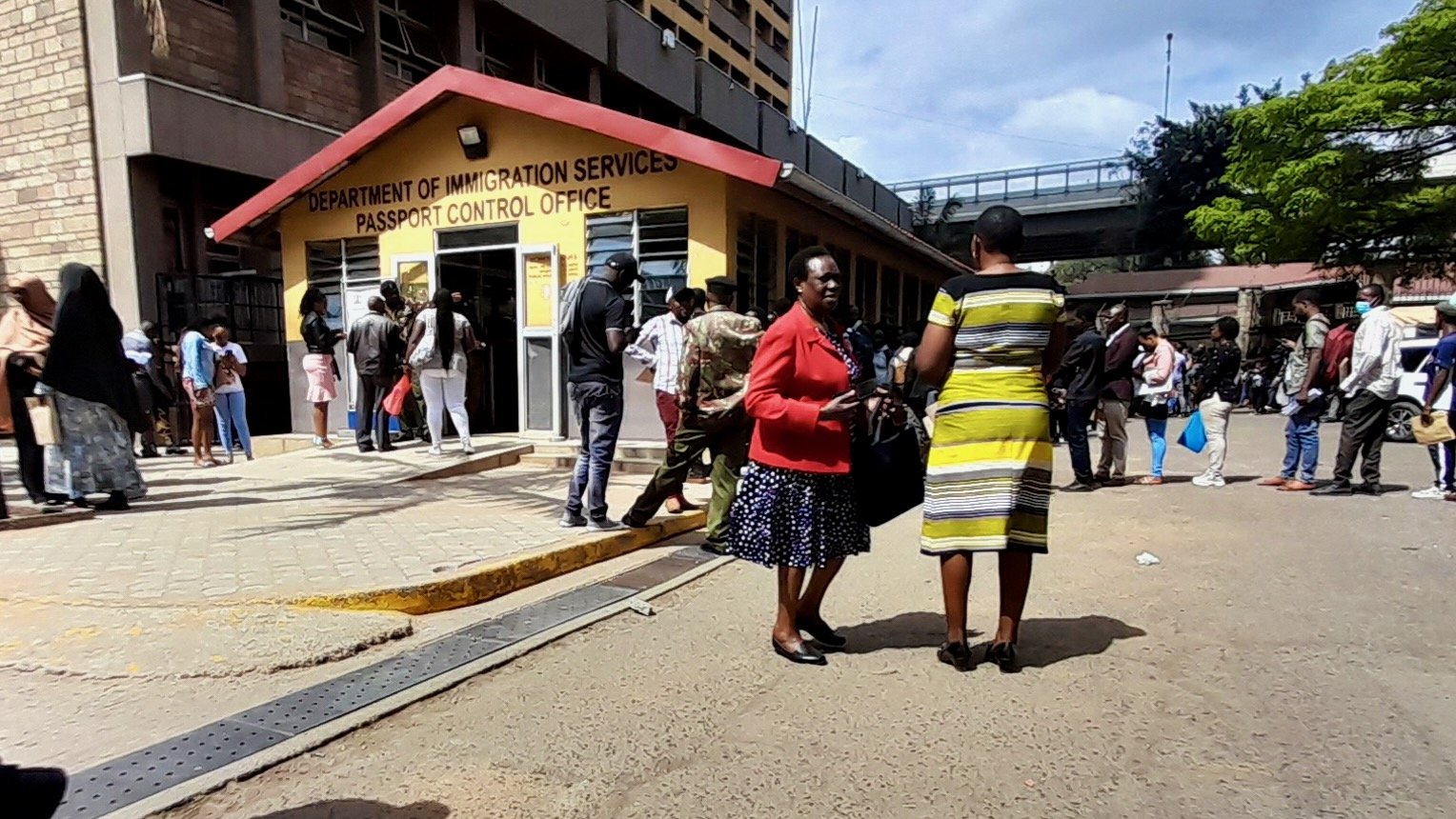 L'image montre une scène animée à l'extérieur d'un bureau de contrôle des passeports, identifié comme le "Department of Immigration Services". À l'avant-plan, deux femmes discutent, l'une portant une jupe et un haut noir avec des motifs, et l'autre vêtue d'une robe rayée jaune. L'environnement est urbain, avec des bâtiments en arrière-plan et une foule de personnes qui attendent ou se déplacent, créant une ambiance dynamique. Le ciel est partiellement nuageux, et l'atmosphère semble être celle d'une journée active. Les surfaces sont en béton, ajoutant un aspect concret à l'ensemble de la scène.