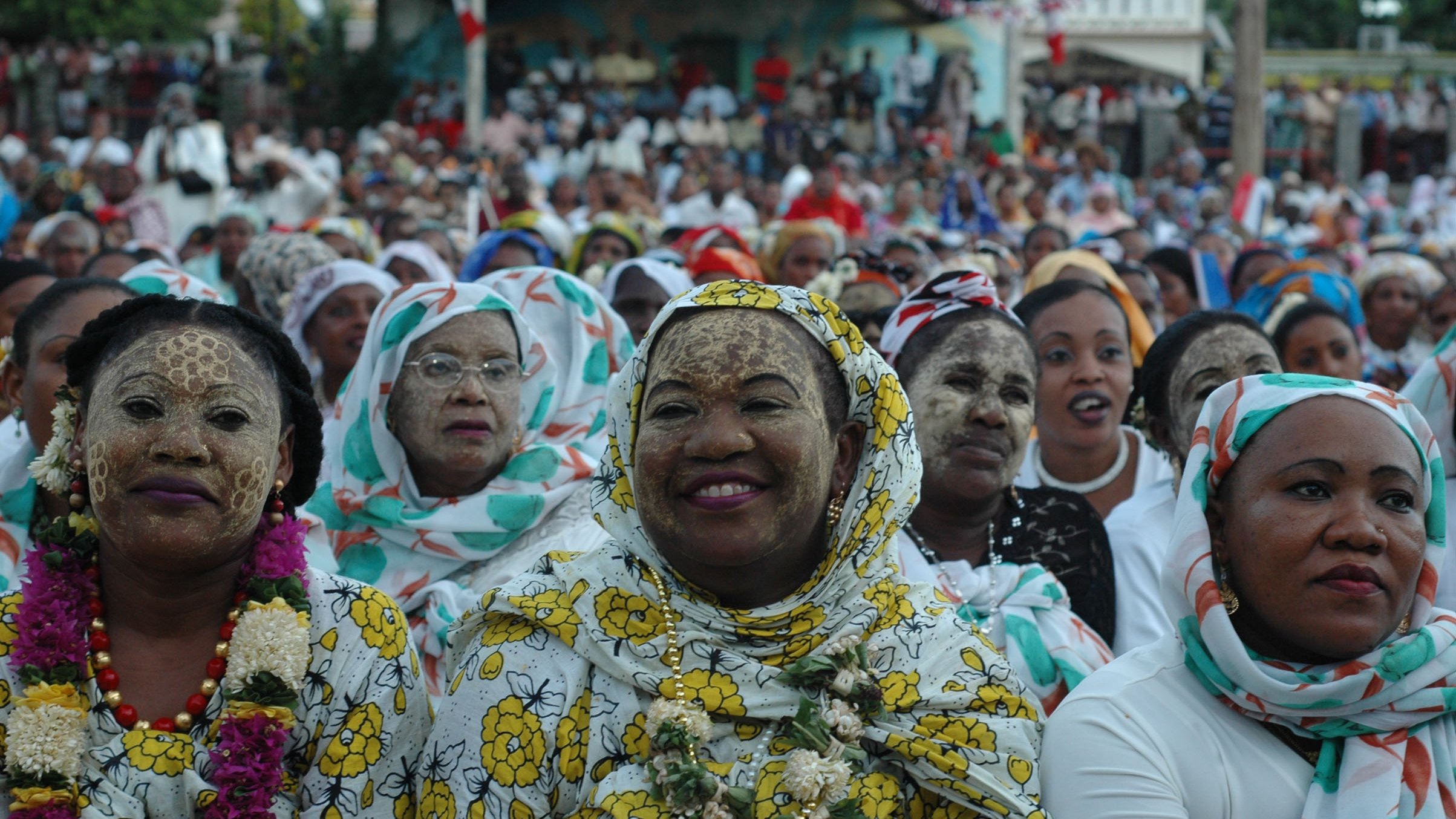 L'image montre un large groupe de personnes réunies pour un événement, probablement festif. Au premier plan, des femmes portent des vêtements colorés et traditionnels, agrémentés de motifs floraux. Certaines ont le visage recouvert de farine ou d'une substance claire, créant une texture particulière. Les femmes semblent joyeuses, souriant et engaged dans l'événement. En arrière-plan, l'assistance s'étend jusqu'à une grande scène, suggérant une atmosphère festive vibrante. Les couleurs et les motifs des vêtements ajoutent une dimension culturelle à l'ensemble de la scène. L'ambiance générale est celle d'une célébration communautaire, remplie de vie et de convivialité.