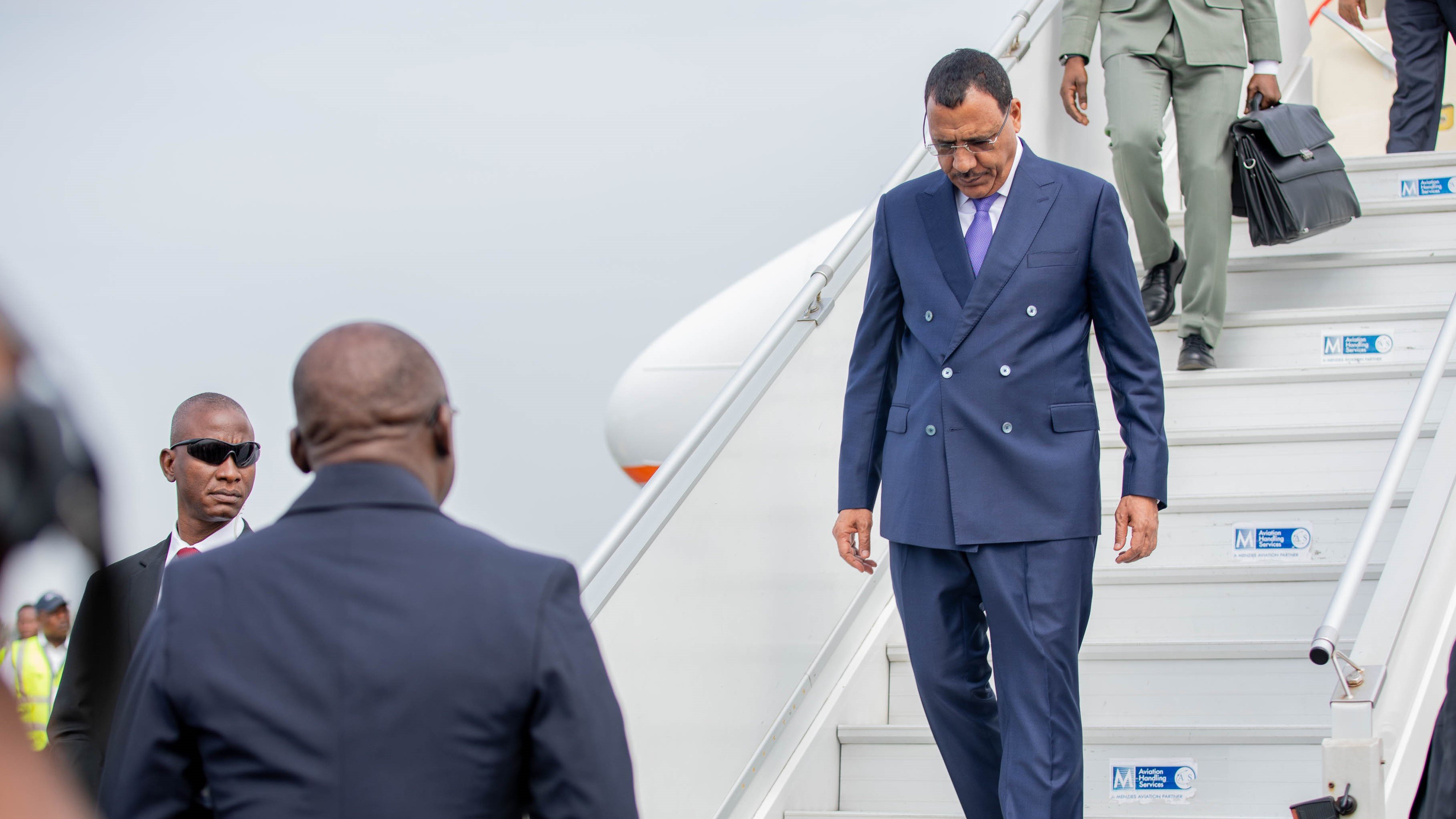 L'image montre un homme qui descend des marches d'un avion. Il porte un costume bleu foncé et des lunettes de soleil. Son expression semble concentrée tandis qu'il s'avance avec assurance. À côté de lui, un autre homme en costume noir observe attentivement. En arrière-plan, on aperçoit des membres du personnel de l'aéroport, ainsi que l'avion stationné. L'atmosphère peut sembler formelle et dynamique, suggérant un moment important, vraisemblablement une arrivée ou une sortie officielle.