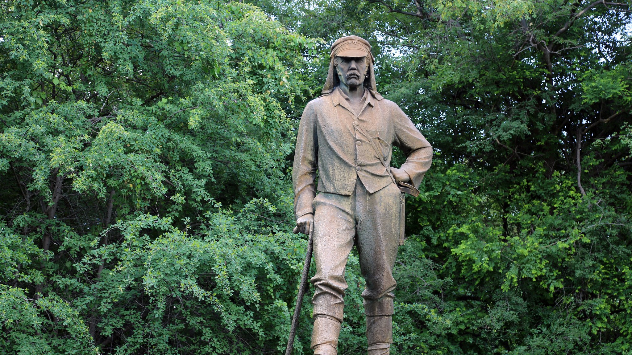 L'image montre une statue en bronze d'un homme qui se tient debout, avec une posture confiante. Il porte des vêtements de style explorateur du XIXe siècle, incluant une chemise à manches longues et un pantalon. Son regard est déterminé et il a une canne à la main, suggérant qu'il pourrait être un aventurier ou un explorateur. La statue est placée sur un socle avec une inscription, et elle est entourée d'une végétation dense, avec des arbres verts en arrière-plan, créant un contraste entre la statue et la nature environnante. L'atmosphère est calme et sereine, reflétant peut-être le respect pour cette figure historique.