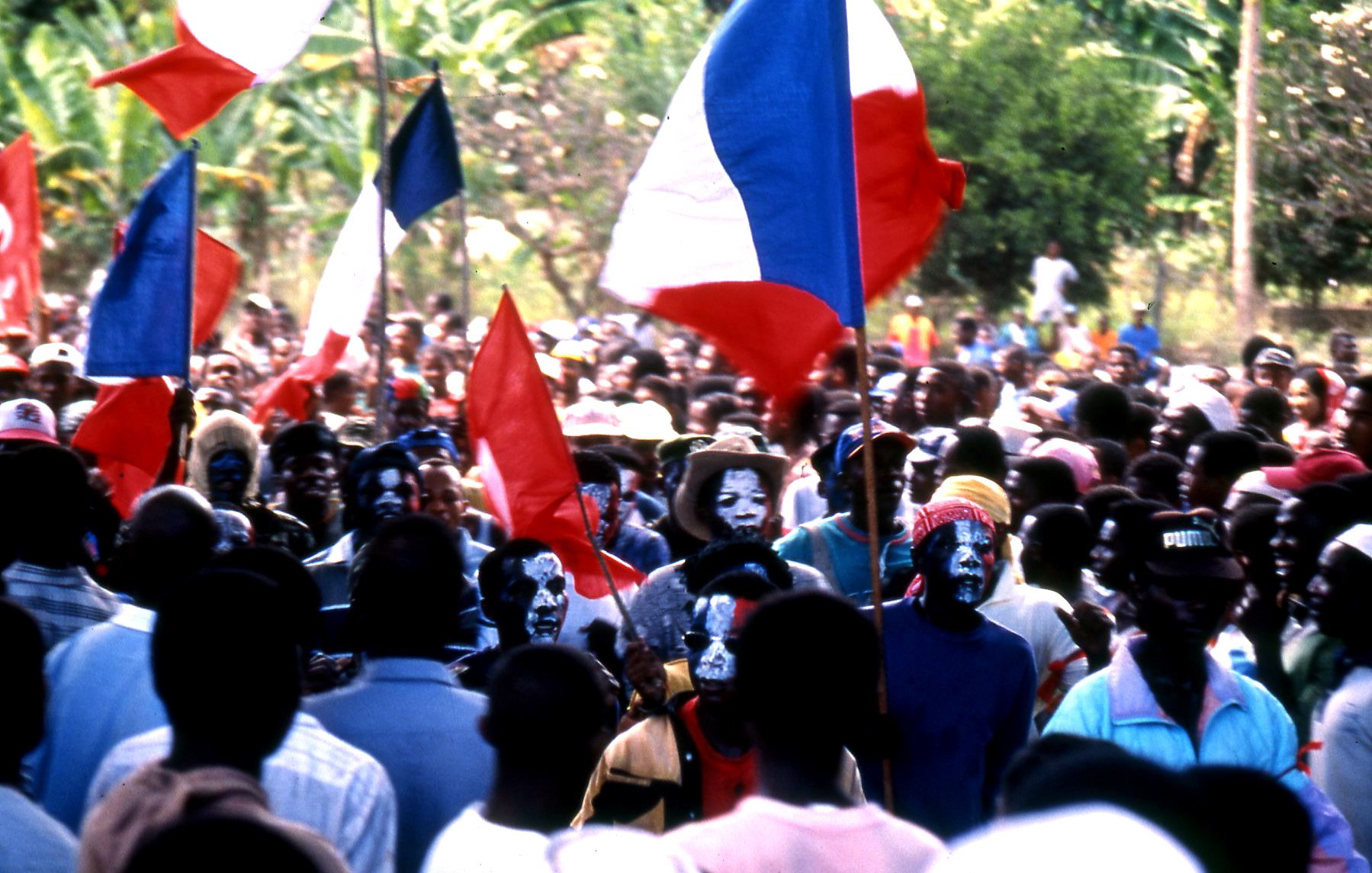 L'image représente une foule dense lors d'un événement, probablement un rassemblement ou une manifestation. Au premier plan, on peut voir des personnes brandissant des drapeaux français, avec des couleurs bleu, blanc et rouge. Certains membres de la foule portent des maquillages colorés sur le visage, ajoutant une dimension festive ou militante à la scène. Les visages sont à la fois heureux et engagés, témoignant d'une ambiance dynamique. En arrière-plan, des arbres et de la verdure suggèrent que l'événement se déroule dans un espace extérieur, peut-être dans un endroit tropical ou rural. L'ensemble dégage une forte impression de solidarité et de passion collective.