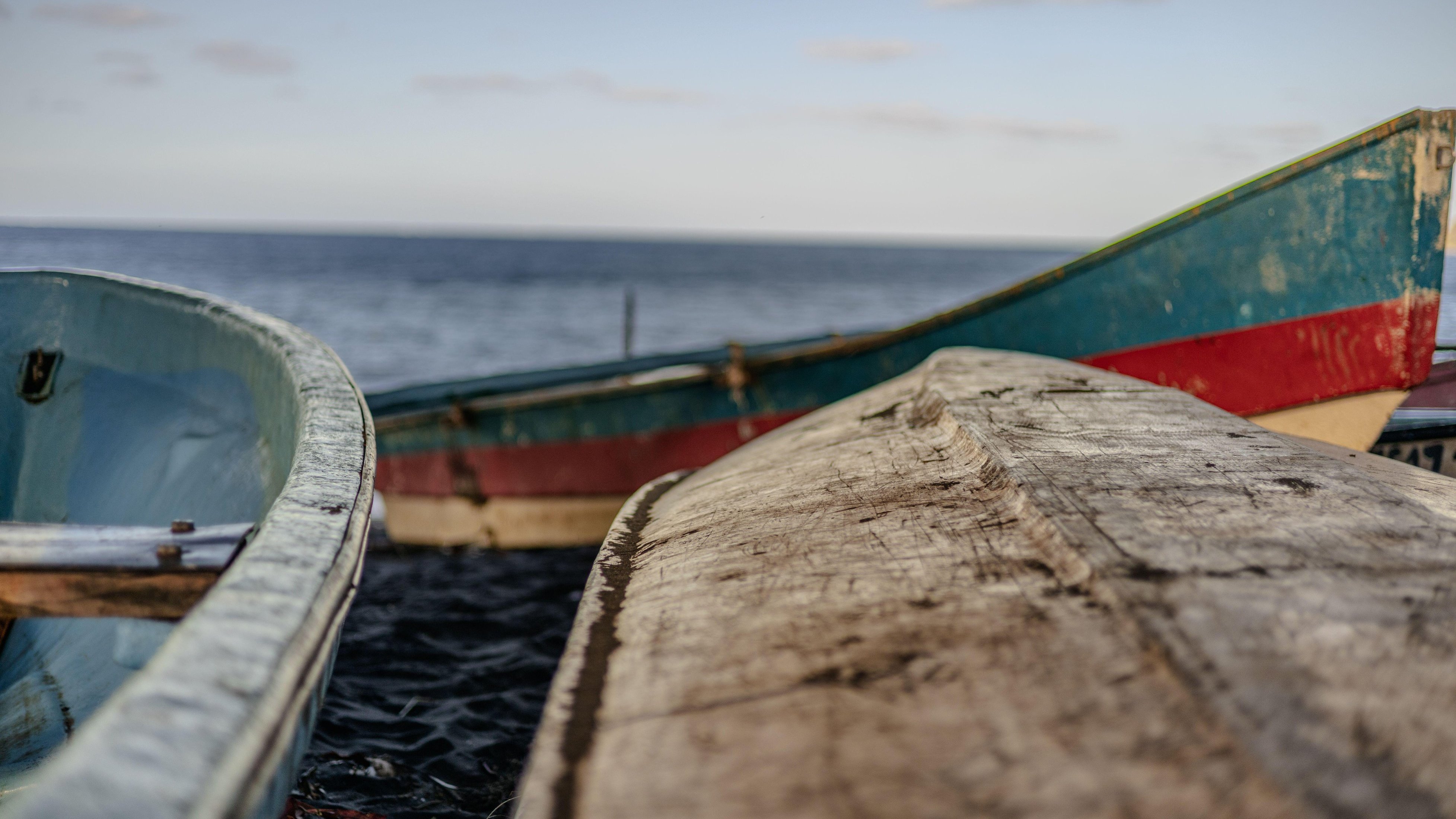 Dans cette image, vous pouvez imaginer un décor paisible au bord de l'eau. Deux bateaux en bois, de designs simples, sont amarrés près d'une plage. Le premier bateau a une structure légèrement arrondie avec une peinture bleu clair sur les côtés, tandis que le second, plus allongé, présente des nuances de bleu et de rouge. Les bateaux sont usés, témoignant de leur utilisation fréquente. En arrière-plan, l'eau s'étend à l'horizon, calme et réfléchissante, et le ciel est dégagé avec quelques nuages légers. L'ambiance est tranquille et maritime, invitant à la contemplation.