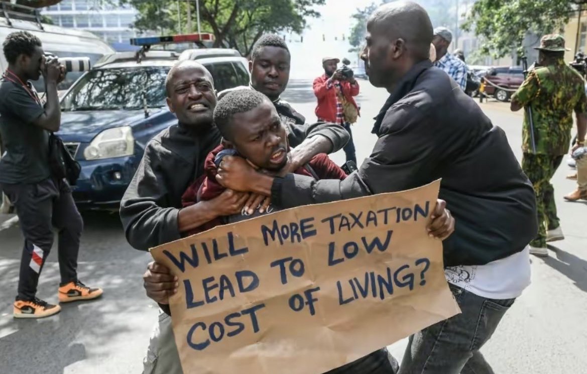 L'image montre une scène de protestation urbaine. Au premier plan, deux hommes sont engagés dans une altercation. L'un d'eux semble essayer de saisir l'autre qui tient une pancarte. Sur cette pancarte, on peut lire une question écrite en lettres noires : "WILL MORE TAXATION LEAD TO LOW COST OF LIVING?" (Plus de taxation conduira-t-elle à un coût de la vie plus bas ?). En arrière-plan, on aperçoit d'autres personnes, certaines les regardent, tandis que d'autres semblent prendre des photos. Des véhicules sont garés sur le côté, ajoutant à l'ambiance animée de la scène. Les expressions des hommes montrent des émotions vives, reflétant l'intensité de la situation.