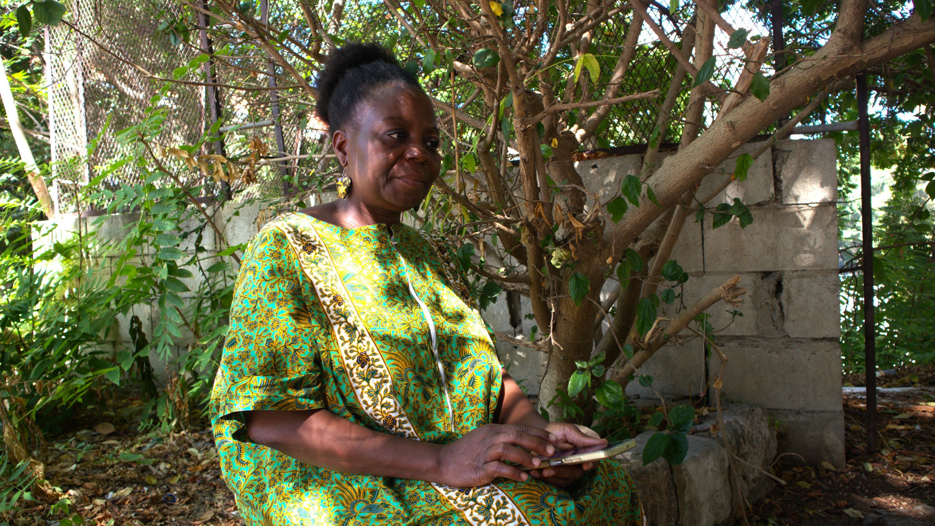 Dans cette image, une femme est assise sous un arbre. Elle porte une robe traditionnelle colorée, dominée par des nuances de vert et des motifs vibrants. Son visage exprime une sérénité et une douce réflexion. À côté d'elle, on peut voir une surface en pierre qui pourrait être un mur. La lumière naturelle filtre à travers les feuillages de l'arbre, créant une atmosphère paisible et intime. Il semble qu'elle tient un objet dans ses mains, probablement un téléphone, et elle est entourée de verdure, ce qui ajoute une touche de vie à la scène.