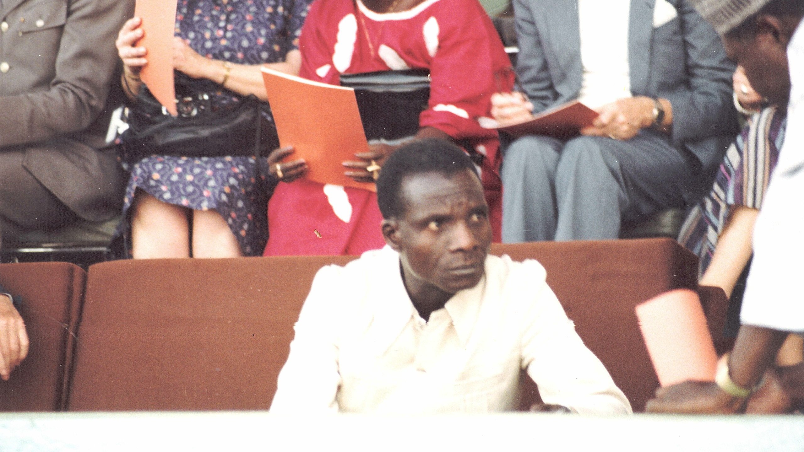 L'image montre une scène d'un événement où un homme en chemise claire est assis au premier plan, semblant sérieux et attentif. À l'arrière-plan, plusieurs personnes discutent, certaines tenant des documents ou des livrets. On aperçoit des vêtements colorés, peut-être d'origine africaine, ce qui ajoute une dimension culturelle à la scène. L'atmosphère générale évoque un rassemblement formel ou une cérémonie.
