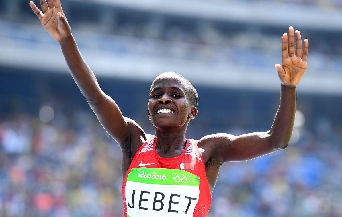 L'image montre une athlète célébrant sa victoire. Elle porte un débardeur rouge avec le nom "JEBET" inscrit dessus, et un logo des Jeux Olympiques sur sa tenue. Ses bras sont levés en signe de triomphe, et elle affiche un large sourire, exprimant la joie et l'excitation de son succès. En arrière-plan, on peut deviner une foule dans un stade, ajoutant à l'atmosphère festive de l'événement. L'éclairage est lumineux, mettant en valeur l'expression de bonheur sur son visage.