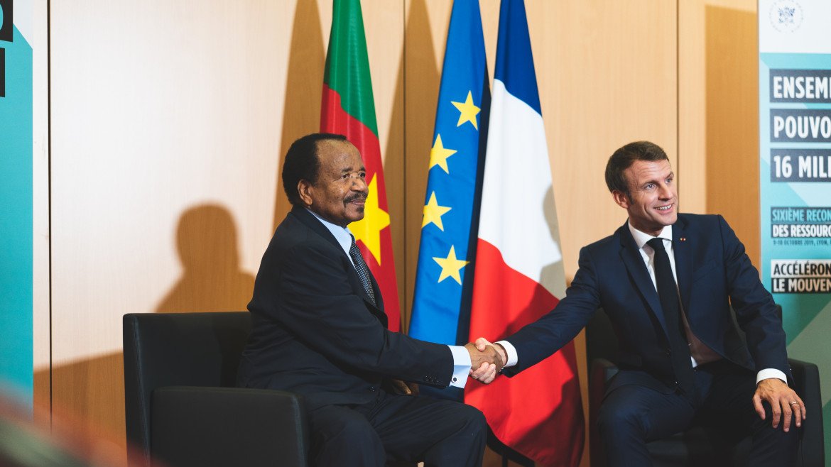 Dans cette image, deux hommes sont assis l'un en face de l'autre, se serrant la main dans un geste formel. L'homme à gauche porte un costume sombre et a une expression souriante. À sa droite, un autre homme, également en costume, affiche un sourire poli. En arrière-plan, on peut noter deux drapeaux : l'un est le drapeau du Cameroun, l'autre le drapeau français et le drapeau de l'Union européenne. L'environnement semble être un espace de réunion officiel, avec des panneaux informatifs visibles. L'atmosphère dégage une impression de cordialité et de coopération.