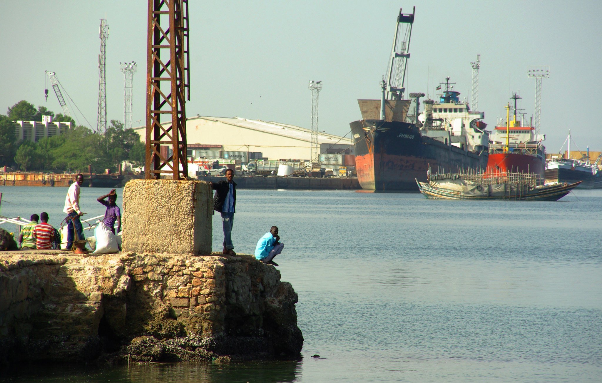 Imaginez un port animé. Au premier plan, il y a un quai en pierre avec plusieurs personnes : certaines sont debout, discutant, tandis que d'autres se penchent sur le bord pour observer l'eau. L'atmosphère est vivante, remplie d'échos de rires et de conversations. À l'arrière-plan, de grands navires sont amarrés, leurs coques visibles au-dessus de l'eau paisible. Des grues et des poteaux élancés se dressent, témoignant des activités portuaires. L'air sent légèrement le sel et le poisson, créant une ambiance maritime authentique.