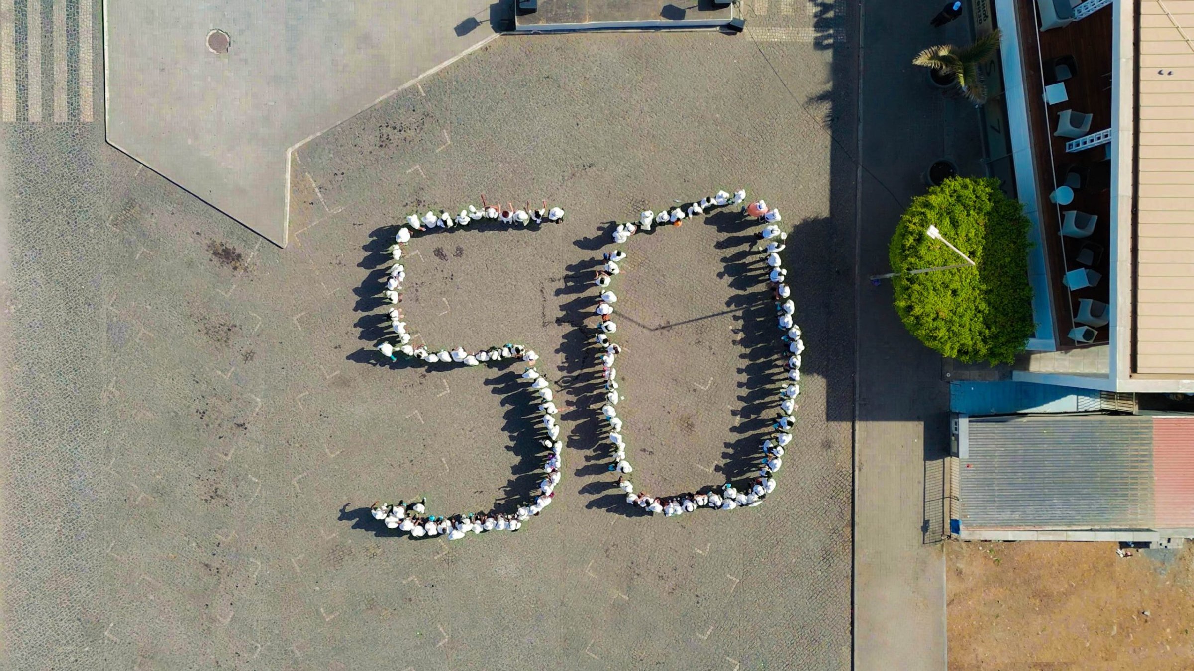 L'image montre une vue aérienne d'une place où des personnes sont réunies pour former le chiffre "50". Les participants sont disposés de manière à créer les deux chiffres avec leurs corps, et ils semblent porter des vêtements clairs, peut-être des t-shirts blancs. Autour, on peut voir des pavés au sol et quelques éléments de paysage urbain, comme des arbres et des bâtiments en arrière-plan. L'ambiance semble conviviale et festive, suggérant que cet événement marque une célébration ou un anniversaire.