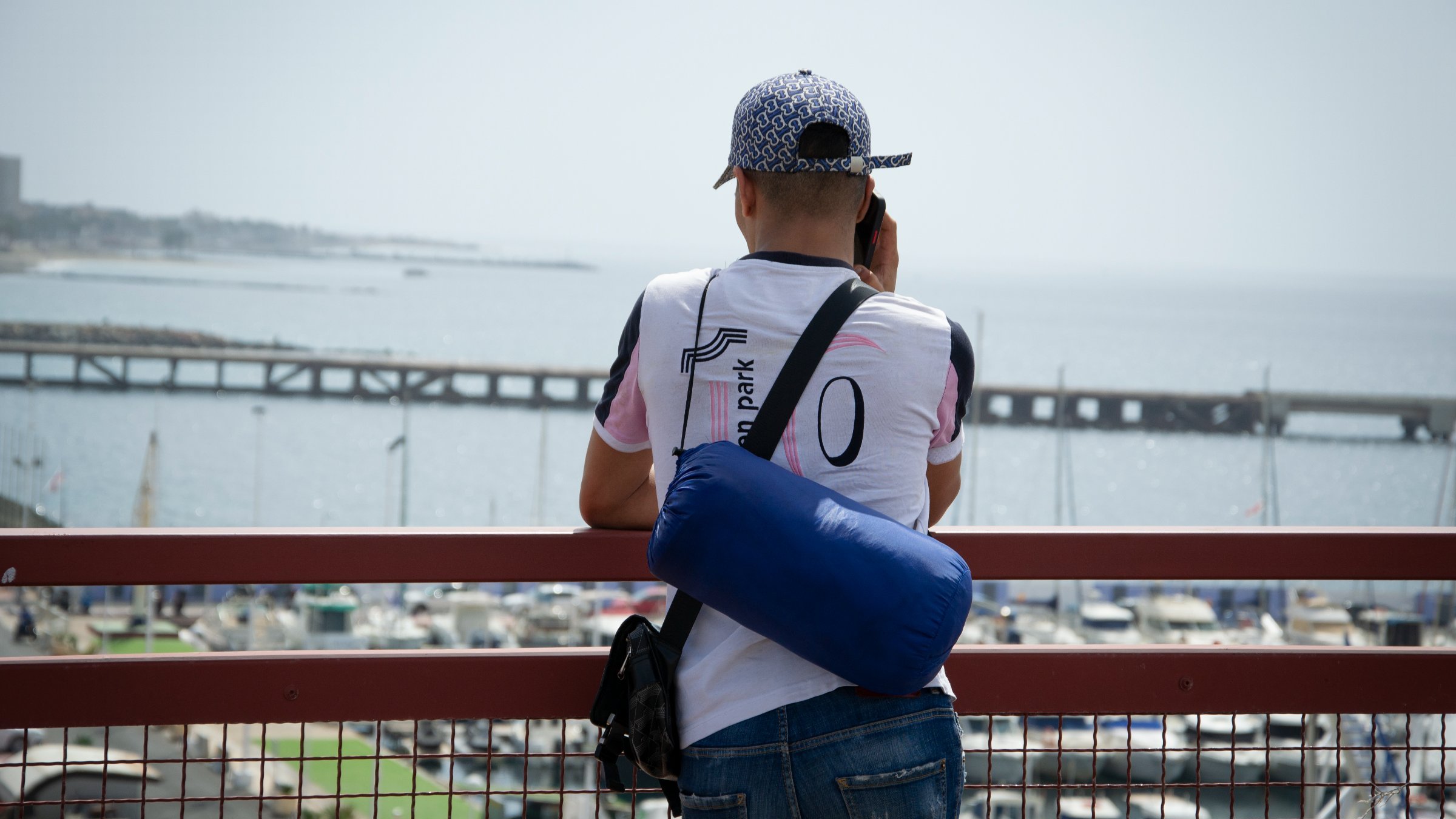 Dans cette image, un homme se tient de dos, regardant au loin vers la mer. Il est vêtu d'un t-shirt blanc avec des accents colorés et porte une casquette à motifs. Sur son épaule, il a un petit sac bleu. L'arrière-plan montre un port avec des bateaux amarrés et une jetée qui s'étend dans l'eau, créant une atmosphère tranquille. Le ciel est clair, suggérant une journée ensoleillée. L'homme semble concentré sur ce qu'il observe, profitant de la vue sur l'horizon maritime.