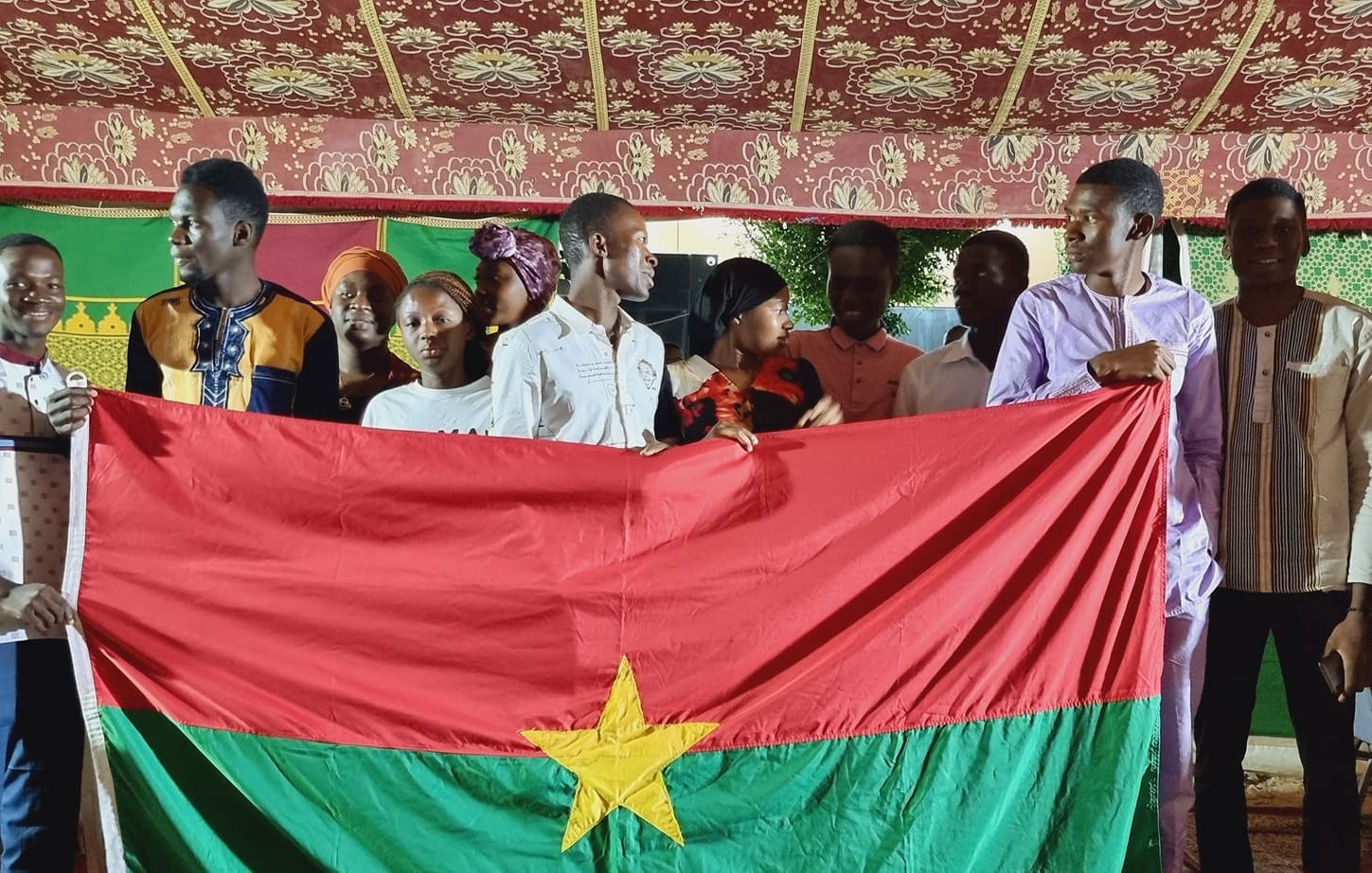 L'image présente un groupe de jeunes personnes rassemblées sous une tente décorée, avec des motifs colorés en arrière-plan. Au centre, ils tiennent une grande drapeau du Burkina Faso, qui est rouge avec une étoile verte au milieu. Les membres du groupe affichent des vêtements variés, certains portant des chemises traditionnelles, et semblent joyeux et solidaires. L'atmosphère est festive, suggérant un événement culturel ou une célébration.