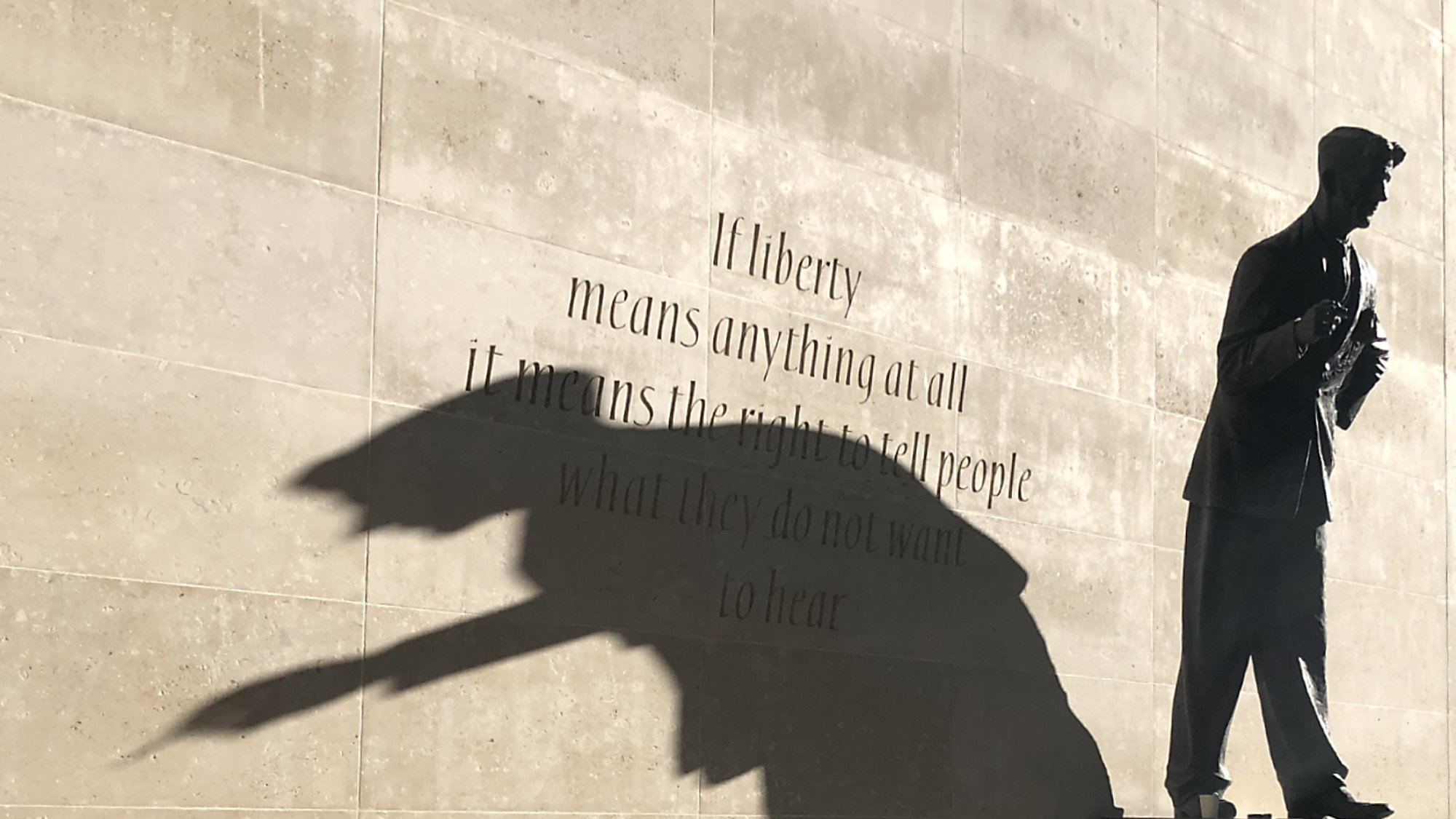 L'image représente une statue d'un homme debout, probablement un auteur ou un penseur, sur un socle. La statue est en bronze, et son ombre projette une forme dynamique sur le mur derrière elle. Ce mur est en pierre lisse, et il porte une citation célèbre qui commence par « If liberty means anything at all... », reflétant des thèmes de liberté et d'expression. L'ensemble crée une atmosphère réfléchie et inspirante, incitant à considérer la profondeur des mots inscrits ainsi que le rôle de la statue.