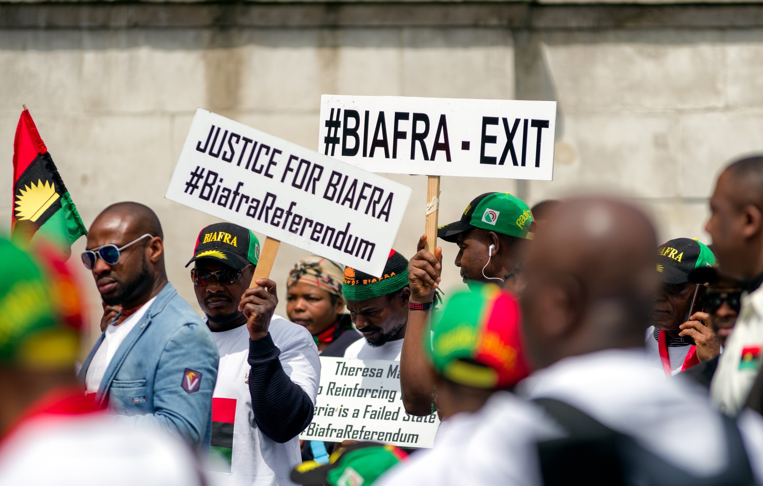 L'image montre un groupe de personnes participant à une manifestation. Elles portent des t-shirts blancs et des casquettes aux couleurs du mouvement Biafra. Des pancartes sont brandies, portant des inscriptions telles que "#BIAFRA - EXIT" et "JUSTICE FOR BIAFRA", ainsi que des hashtags liés au référendum biafrais. L'atmosphère semble dynamique et engagée, reflétant une revendication pour la justice et l'autodétermination.