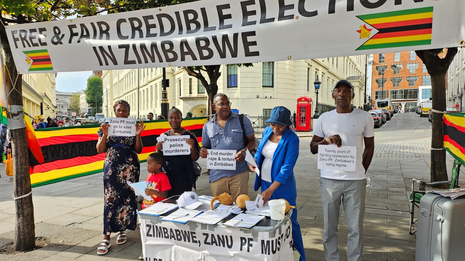 L'image montre un groupe de personnes rassemblées sous une grande banderole qui proclame "Élections libres et équitables au Zimbabwe". Cette banderole est ornée des couleurs du drapeau zimbabwéen, avec du rouge, du jaune, du vert et du noir. Les manifestants tiennent des panneaux avec des messages portant sur la nécessité de respecter les droits humains et d'organiser des élections justes au Zimbabwe. Sur la table devant eux, il y a des brochures et des documents qui semblent orientés vers la sensibilisation et l'information sur la situation politique. L'environnement est urbain, avec des bâtiments en arrière-plan et des passants qui s'intéressent à la manifestation.