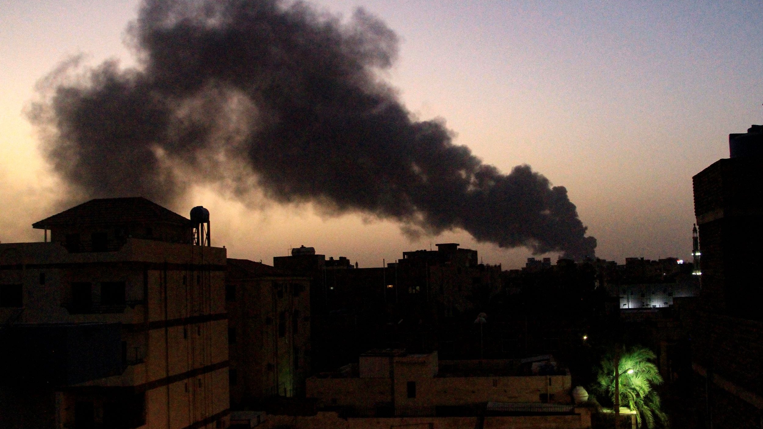 L'image montre un paysage urbain au crépuscule, où le ciel est teinté de nuances de bleu et d'orange. En arrière-plan, une épaisse colonne de fumée noire s'élève, suggérant une source d'incendie. Les silhouettes de bâtiments se dessinent en avant-plan, avec des structures variées et quelques lumières allumées. À droite, on aperçoit un palmier, ajoutant une touche de verdure à ce paysage sombre et chargé d'une ambiance d'urgence.