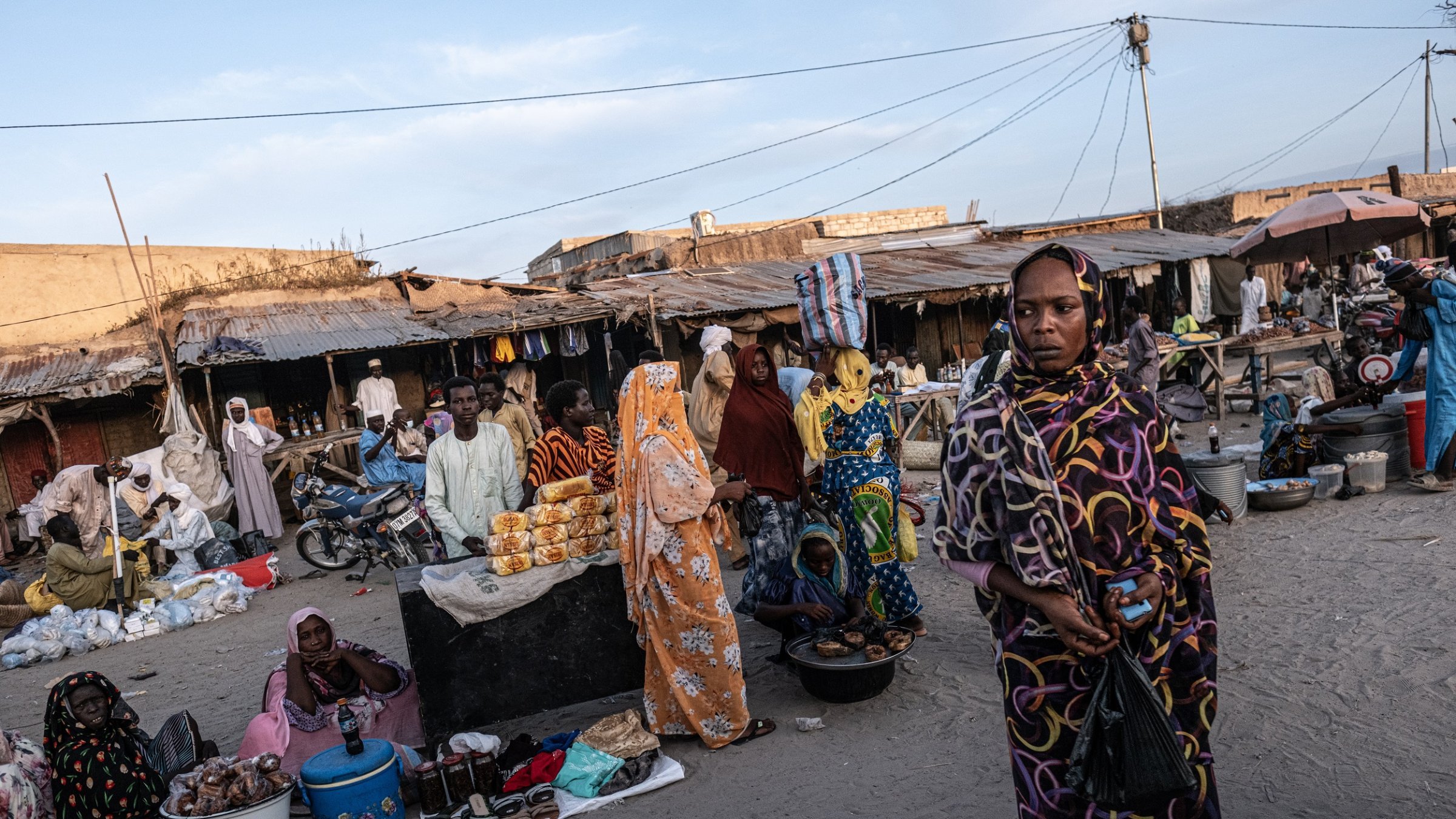 Dans cette scène animée d'un marché, on peut ressentir une atmosphère vibrante. Le sol est recouvert de sable, et plusieurs stands sont disposés tout autour. Des gens sont rassemblés, certains vêtus de vêtements colorés, offrant divers produits alimentaires, tels que des fruits et des pâtisseries. Les femmes, en particulier, attirent l'attention avec leurs robes chatoyantes et leurs voiles. On peut aussi entendre des murmures de conversations, des rires et le bruit de petits véhicules, comme des motos, qui se déplacent à travers le marché. L'air est imprégné d'odeurs variées de nourriture épicée et de douceur, créant une expérience sensorielle unique.