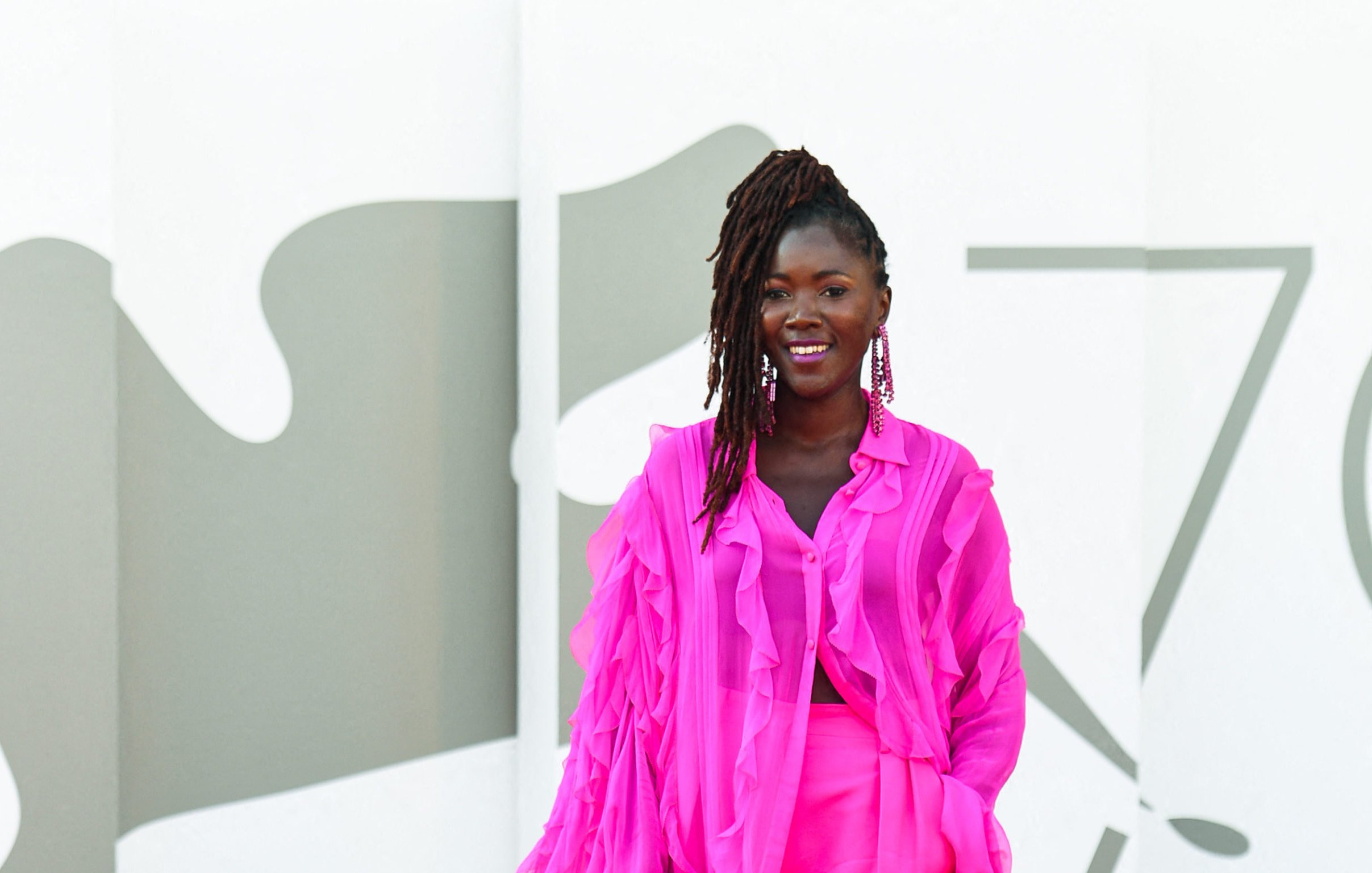 L'image montre une femme debout sur un fond minimaliste. Elle porte une tenue éclatante de couleur rose vif, composée d'une blouse légère aux manches froncées et d'une jupe assortie. Son sourire est radieux et elle affichent une attitude pleine de confiance. Ses cheveux sont coiffés en dreadlocks, et elle porte de grandes boucles d'oreilles qui ajoutent une touche élégante à son look. Le fond est composé de formes géométriques en tons neutres, mettant en valeur la vivacité de sa tenue.