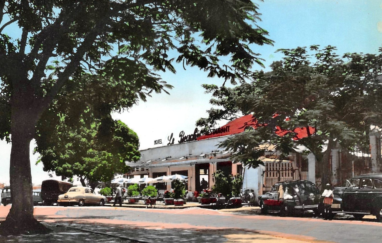 L'image montre une scène d'un restaurant, probablement située dans un quartier ensoleillé. À l'avant, on aperçoit une route bordée d'arbres dont les feuilles offrent un peu d'ombre. Le bâtiment principal, assez spacieux, possède un toit rouge et est marqué par l'inscription "Le Restaurant" en lettres blanches. Plusieurs voitures anciennes, de différentes couleurs, sont garées près de l'établissement. Des personnes flânent autour, créant une atmosphère de vie et de convivialité. En arrière-plan, le ciel est bleu, ce qui ajoute une touche de chaleur et de clarté à l'ensemble de l'image.