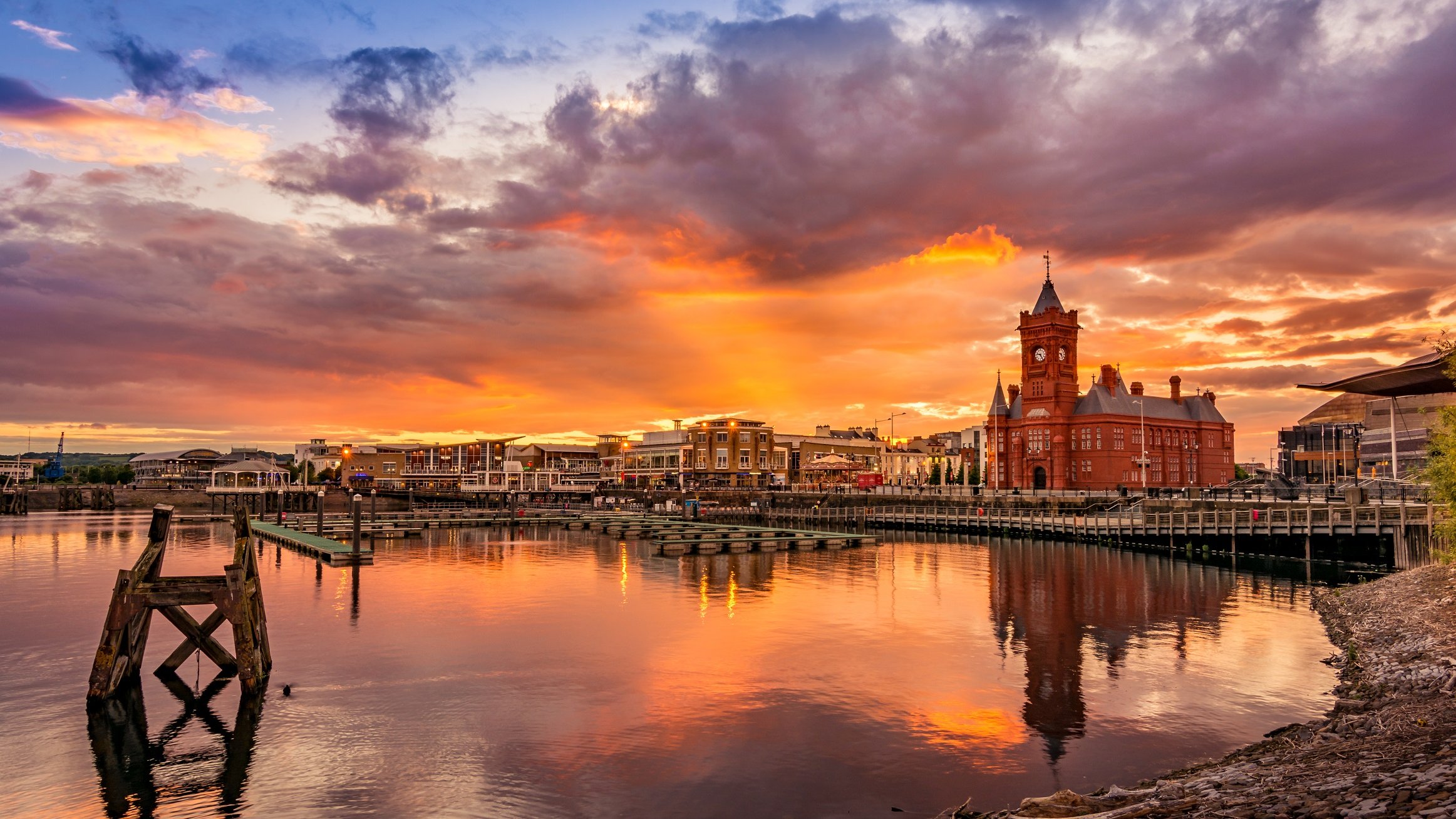 Cette image représente un paysage de bord de mer au coucher du soleil. À l'horizon, un ciel splendide est illuminé par des teintes enflammées de rouge, orange et violet. Le soleil déclinant éclaire les nuages, créant une atmosphère magique. Au premier plan, l'eau calme d'une marina reflète ces couleurs vives, tandis que plusieurs quais et petits bateaux ajoutent à la scène paisible. Sur la rive, un bâtiment architectural distinctif de couleur rouge, avec une tour pointue, attire l'attention, tandis que des arbres et des éléments naturels encadrent le paysage, offrant une ambiance sereine et accueillante.