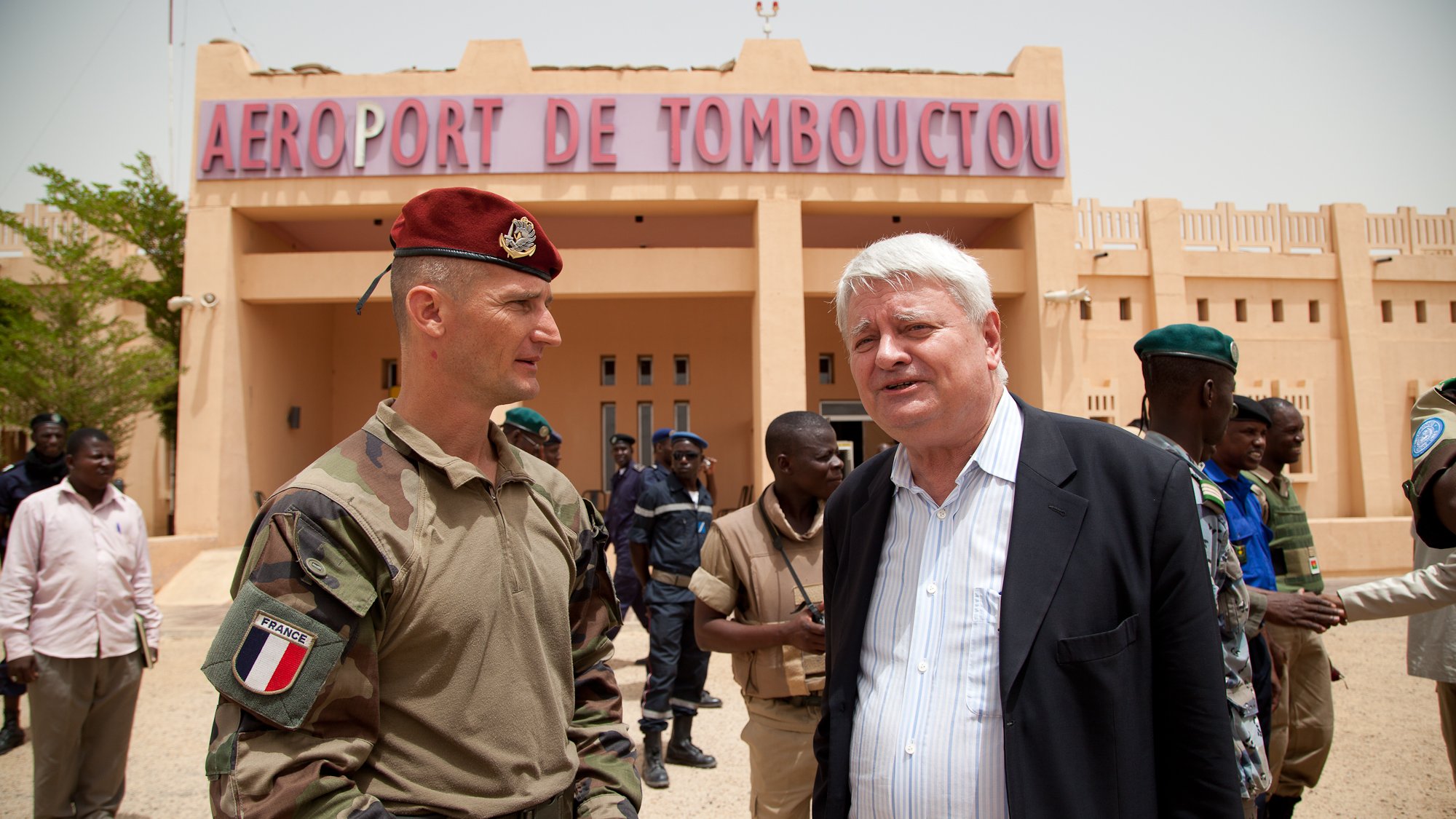 L'image montre une scène à l'aéroport de Tombouctou, avec un bâtiment beige en arrière-plan portant le nom "AÉROPORT DE TOMBOUCTOU" en lettres rouges. Au premier plan, deux hommes sont en conversation. L'un est en uniforme militaire, portant un béret rouge, tandis que l'autre est habillé en costume. Autour d'eux, plusieurs personnes en uniforme, probablement des militaires et des policiers, sont présentes, créant une atmosphère chargée d'activités. Le ciel est clair et le paysage indique une région chaude, avec un sol désertique.