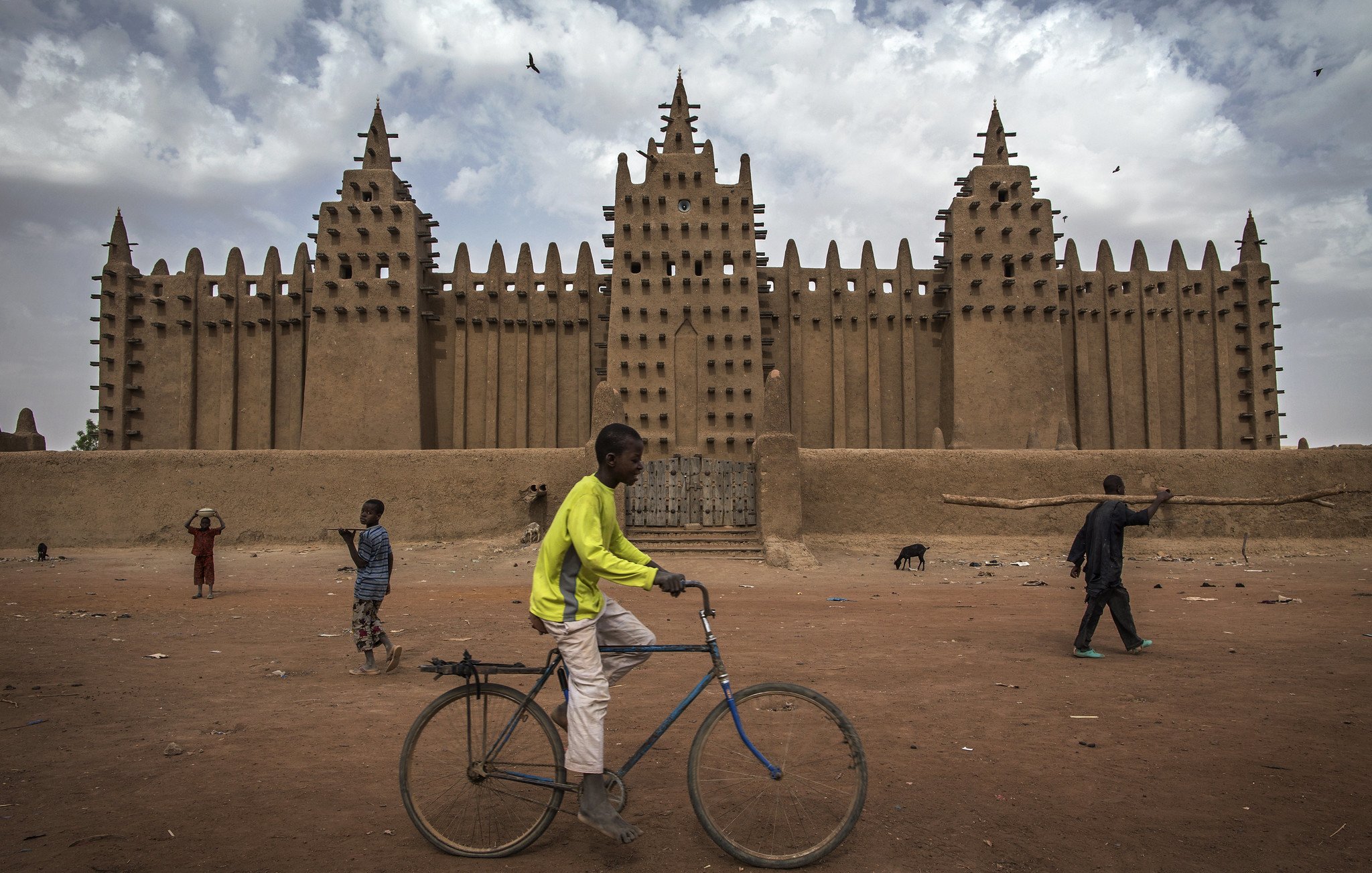 L'image montre une grande mosquée en banco, typique de l'architecture soudanaise, avec des murs en terre crue qui s'élèvent vers le ciel. Sa façade est ornée de nombreuses pointes et créneaux, formant un motif impressionnant. Au premier plan, un jeune garçon traverse la scène à bicyclette, tandis que d'autres enfants jouent et se déplacent dans l'espace. Le sol est poussiéreux, et le climat semble chaud, avec des nuages ici et là dans le ciel. L'ambiance est vivante, illustrant une scène animée de la vie quotidienne.