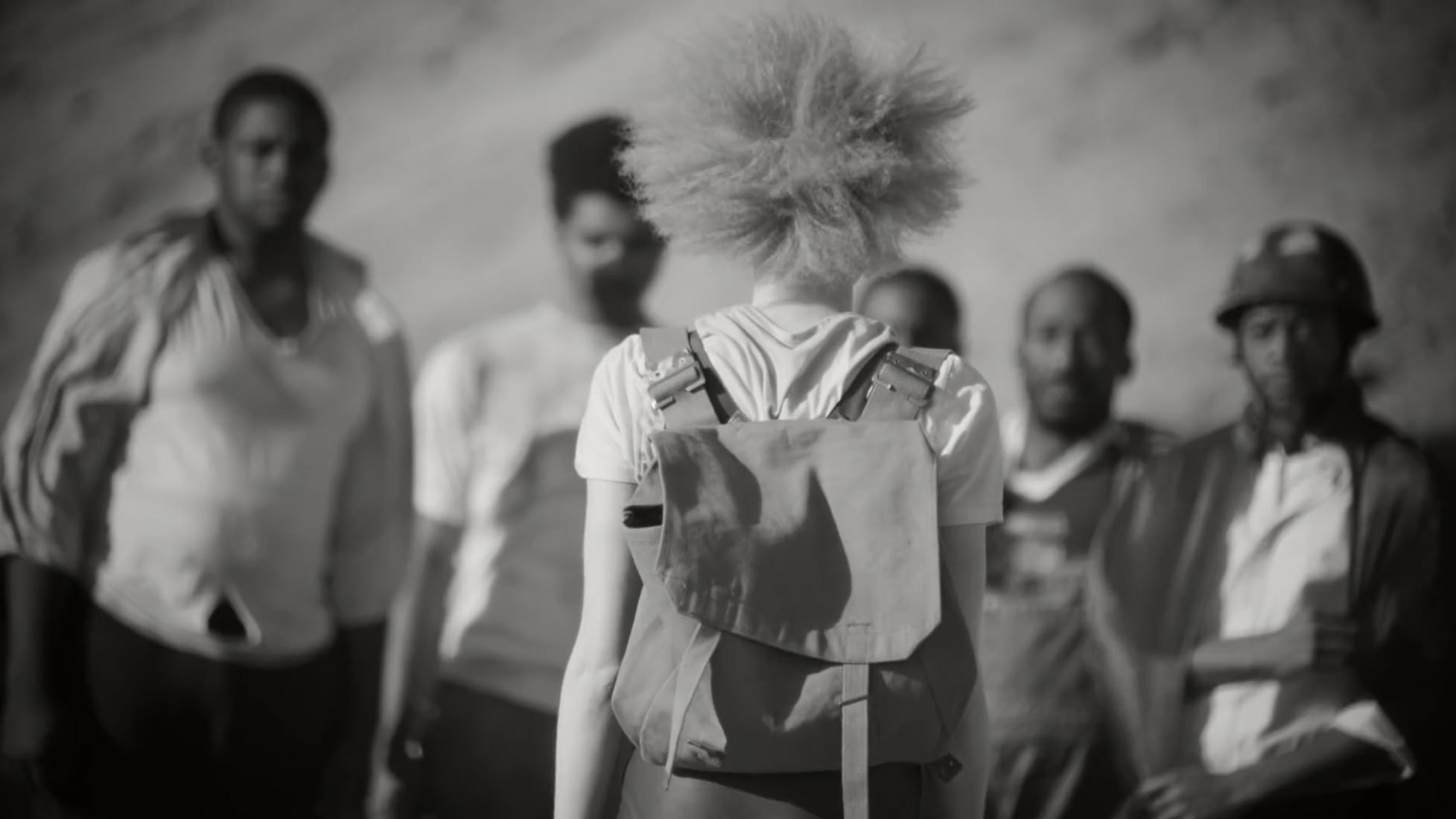L'image présente un groupe de personnes, avec au premier plan une personne aux cheveux crépus et blonds, vue de dos. Elle porte un sac à dos. L'arrière-plan est flou, mais on devine des montagnes ou des collines. Les autres individus, qui se tiennent plus loin, ont des expressions faciales sérieuses et semblent réunis dans une conversation ou une interaction. L'atmosphère de l'image est en noir et blanc, ce qui accentue les contrastes entre les différentes textures et les attitudes des personnages.