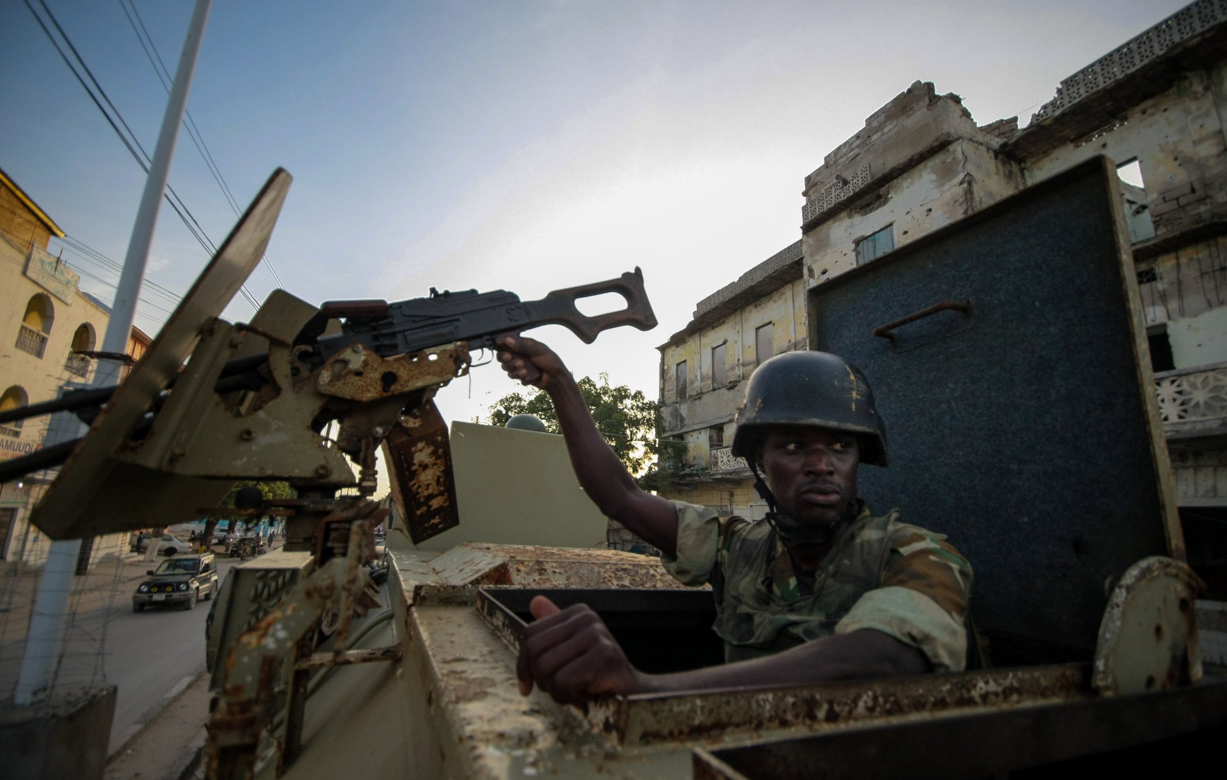 L'image montre un soldat assis dans un véhicule militaire. Il est vêtu d'un uniforme camouflage et porte un casque. Le soldat tient un fusil d'assaut, que l'on peut imaginer prêt à être utilisé. En arrière-plan, on aperçoit des bâtiments en ruine, suggérant que l'environnement est marqué par des signes de conflit. La lumière est douce, indiquant peut-être un lever ou un coucher de soleil. L'atmosphère dégage un sentiment de tension et de vigilance dans un cadre urbain dégradé.