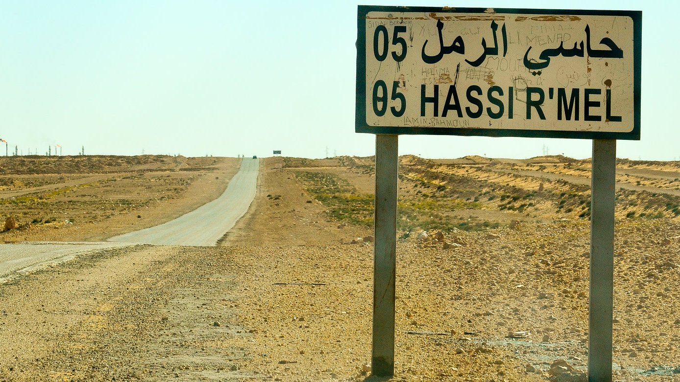 L'image montre une route désertique dans un paysage aride. À droite, un grand panneau de signalisation indique "Hassi R'Mel" en arabe et en français, avec le numéro 05. En arrière-plan, on aperçoit des structures industrielles, dont une cheminée qui dégage de la fumée, suggérant l'activité d'extraction de ressources. L'environnement est principalement constitué de terres sèches et peu de végétation, illustrant une atmosphère isolée et stérile. Le ciel est clair, et la chaleur du jour est palpable.