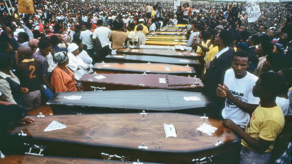 L'image montre une vaste foule rassemblée en extérieur, entourant une série de cercueils alignés. Les cercueils, de différentes couleurs et styles, sont disposés côte à côte, symbolisant une cérémonie funèbre. Les personnes présentes semblent nombreuses et engagées, certaines portent des vêtements de deuil, tandis que d'autres affichent des expressions de tristesse ou de solidarité. L'atmosphère est empreinte de respect et de recueillement, évoquant une perte collective. Au fond, des pancartes sont visibles, ajoutant une dimension politique ou sociale à l'événement. Le ciel est nuageux, renforçant le caractère solennel de la scène.