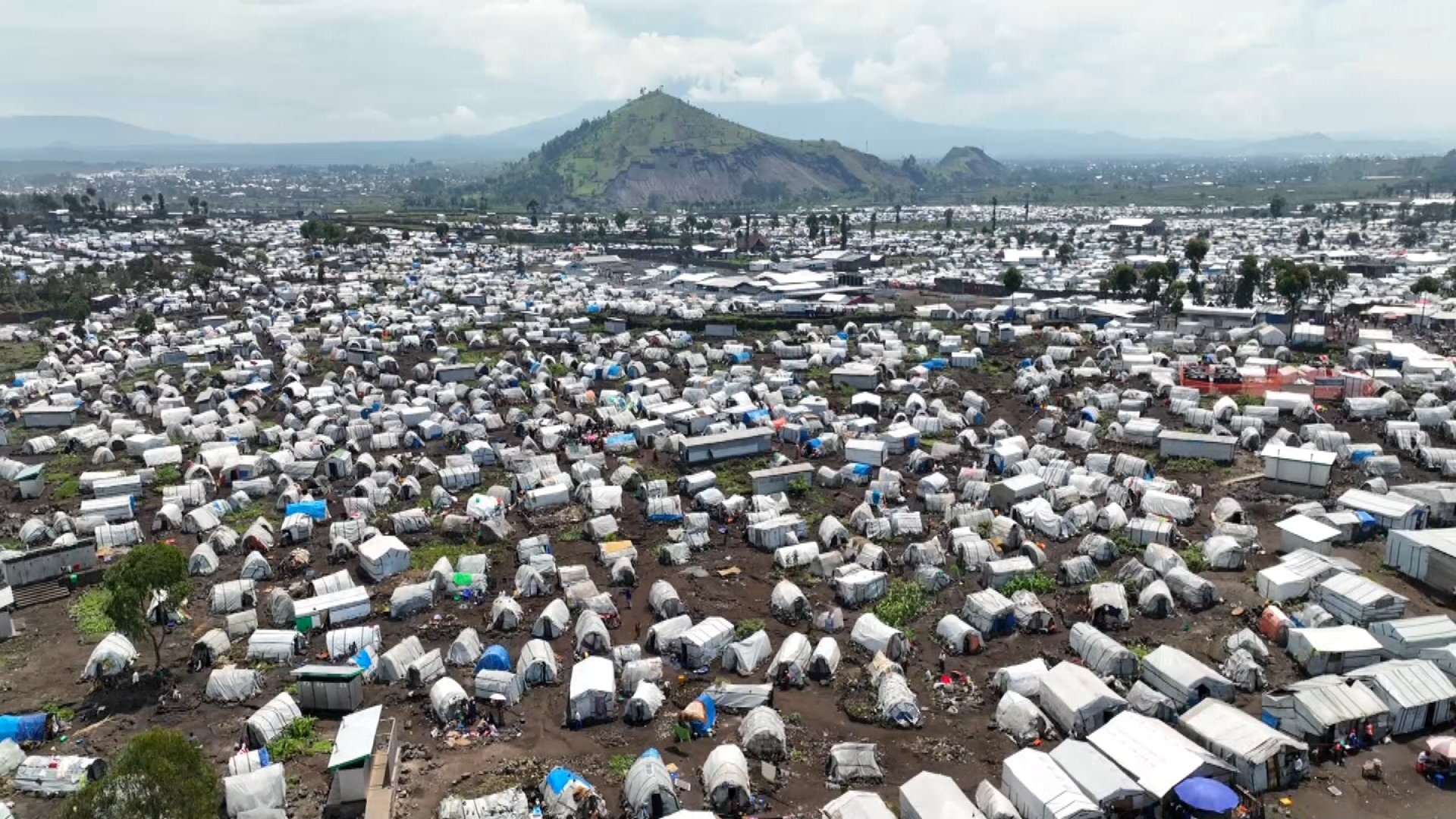 L'image montre un vaste camp de tentes, probablement un site de secours ou un camp de réfugiés, s'étendant à perte de vue. Les tentes sont de couleurs multiples, avec des toits en toile qui varient du blanc au bleu, et certaines sont en désordre avec des morceaux de tissu et de plastique. Le terrain est accidenté, avec des traces d'usage et de la terre visible entre les tentes. En arrière-plan, on aperçoit une montagne verdoyante qui domine le paysage, ajoutant un contraste entre la nature et l'afflux humain. À distance, on distingue des constructions urbaines, des routes et d'autres installations, suggérant la proximité d'une ville. L'ensemble de la scène véhicule une impression de densité et de survie collective, caractéristique des situations d'urgence humanitaire.