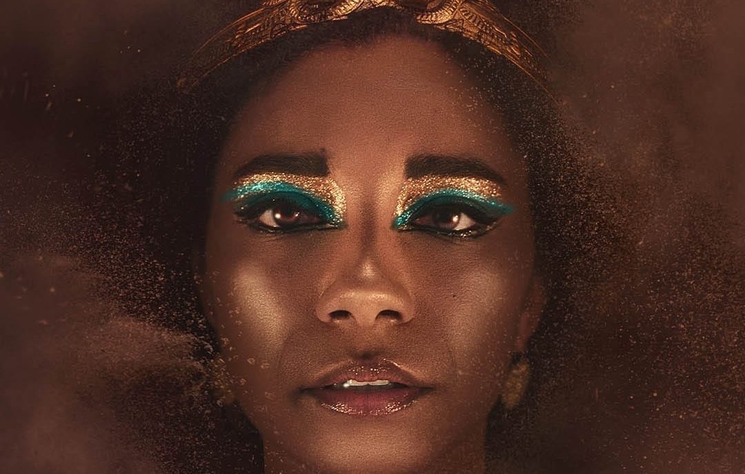 L'image montre un portrait d'une femme avec une peau sombre et un regard intense. Elle porte une couronne dorée ornée de serpents. Son maquillage se distingue par des teintes vibrantes de turquoise autour des yeux, ajoutant un contraste frappant à son visage. La lumière douce crée une ambiance mystérieuse, tandis qu'un brouillard léger autour d'elle donne une impression d'éthéré. L'ensemble dégage une aura de majesté et de puissance.