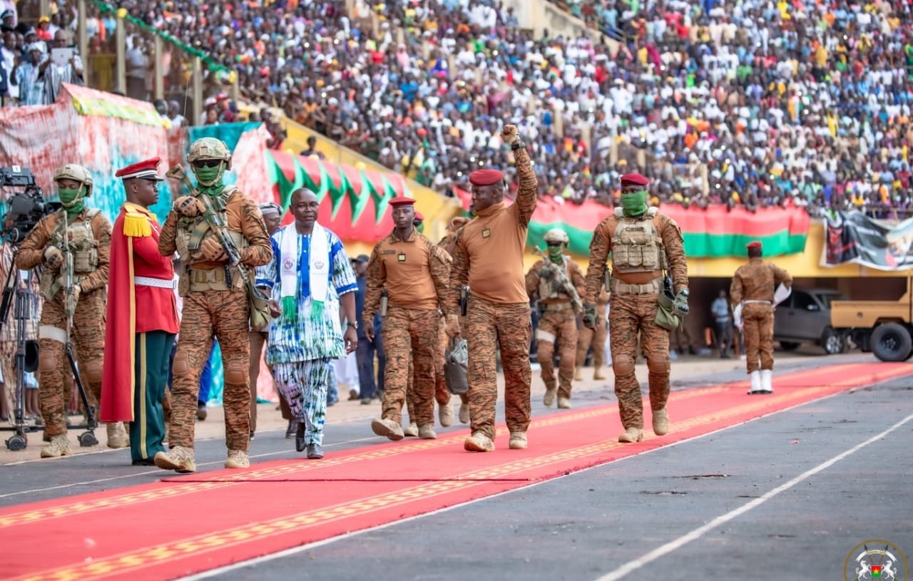 L'image dépeint une scène d'événement public dans un stade. Un grand nombre de personnes est rassemblé dans les gradins, créant une ambiance festive et patriotique. Au premier plan, des militaires en uniforme brun avancent sur un tapis rouge, symbolisant une cérémonie officielle. L'un des militaires, portant une casquette rouge, lève le bras en signe de salutation. À ses côtés, un homme en vêtements traditionnels aux motifs colorés marche avec assurance. La scène est remplie de drapeaux et de banderoles qui ajoutent des touches de couleur à l'événement. L'atmosphère est vibrante, marquée par l'engagement et l'unité.