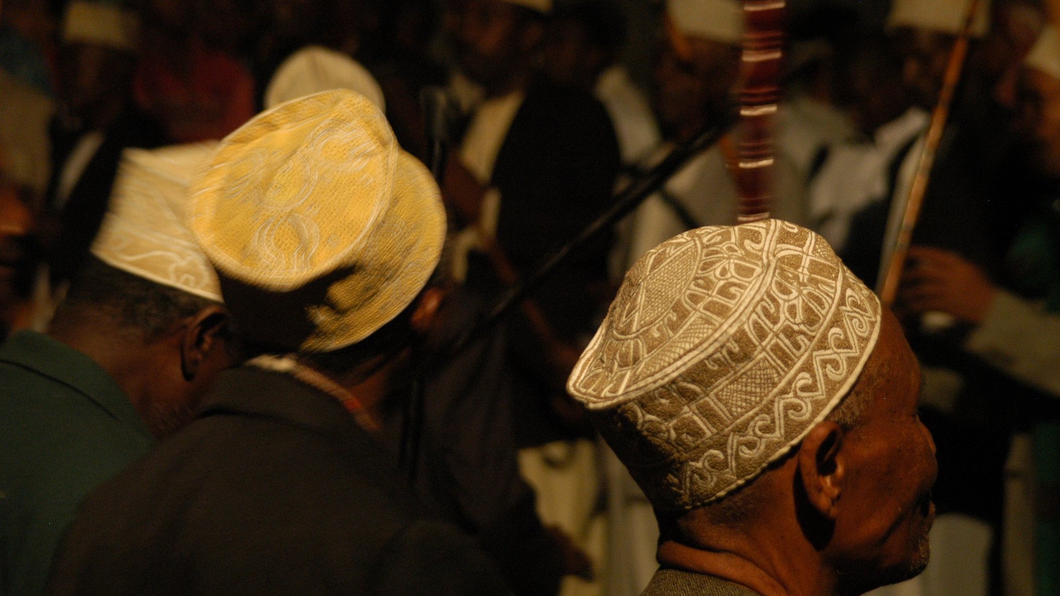 L'image présente un groupe de personnes, principalement des hommes, de profil. Ils portent des chapeaux traditionnels, souvent ornés de motifs délicats, qui varient en couleurs et en designs. En arrière-plan, on peut distinguer d'autres figures et instruments de musique, suggérant une ambiance festive ou rituelle. La lumière est tamisée, créant une atmosphère intime. Les visages des hommes sont flous, mais leur posture et leurs chapeaux ajoutent une touche culturelle à la scène.