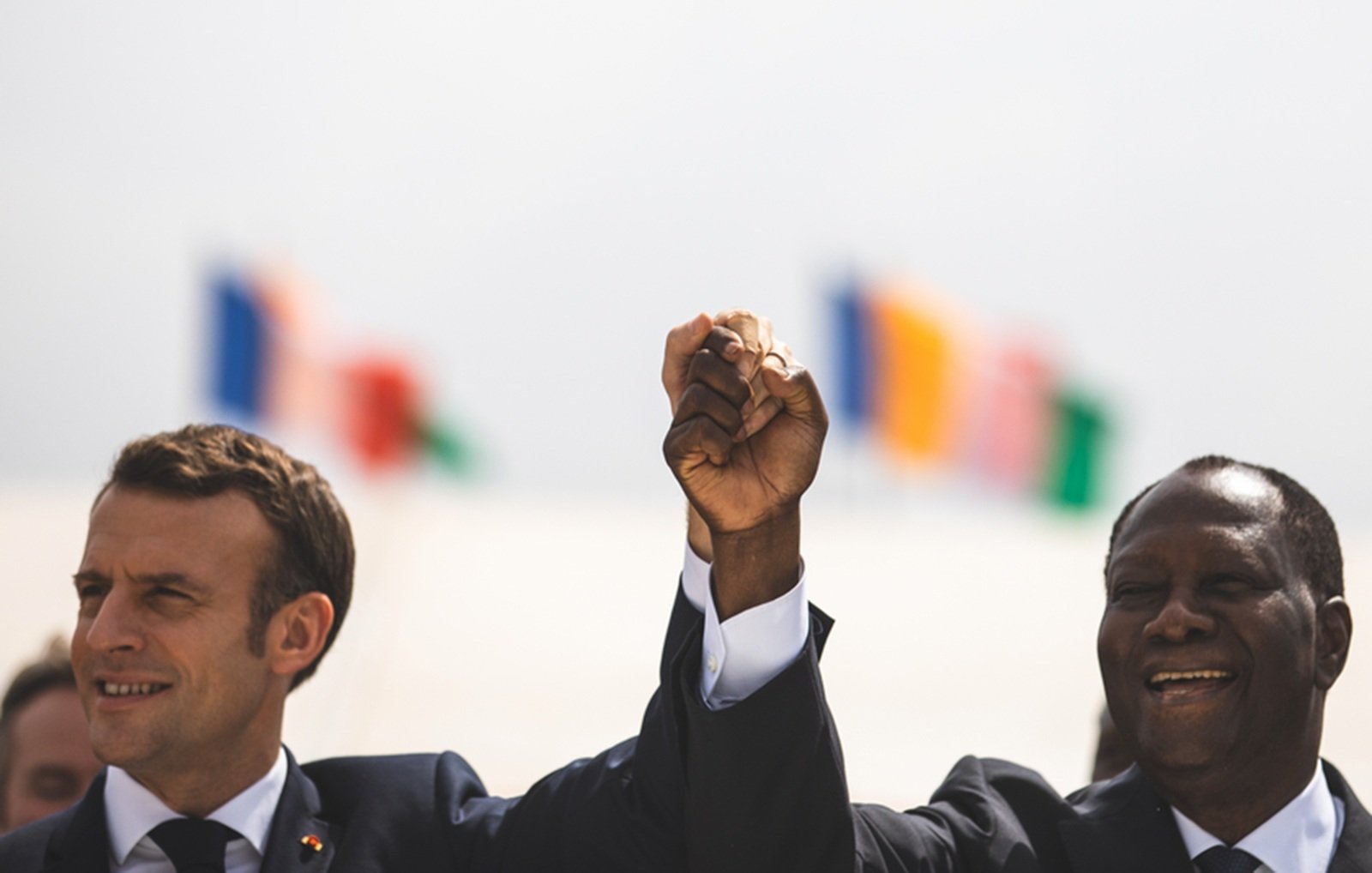 Imaginez une scène marquée par un moment de camaraderie et d'unité. Deux hommes se tiennent côte à côte, levant la main droite en l'air, leurs poings serrés, symbolisant la force et la solidarité. À gauche, on perçoit un homme avec une coupe de cheveux soignée, vêtu d'un costume sombre, tandis que l'homme à droite, également en costume, affiche un grand sourire chaleureux. En arrière-plan, plusieurs drapeaux flottent doucement au vent, représentant différentes nations. L'atmosphère est festive et pleine d'espoir.