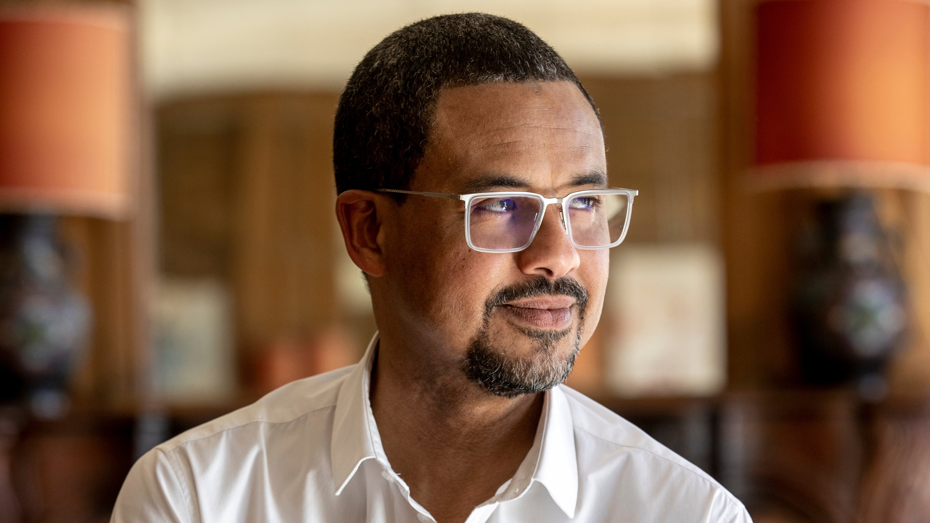 L'image présente un homme d'âge moyen, debout légèrement sur le côté. Il a la peau foncée et porte des lunettes avec une monture claire. Son expression est contemplative, avec un léger sourire aux lèvres. Il porte une chemise blanche, qui contraste avec son teint. En arrière-plan, on peut apercevoir des lampes avec des abat-jour orange et un décor en bois qui donne une ambiance chaleureuse et intime à l'ensemble. La lumière est douce, créant une atmosphère paisible.