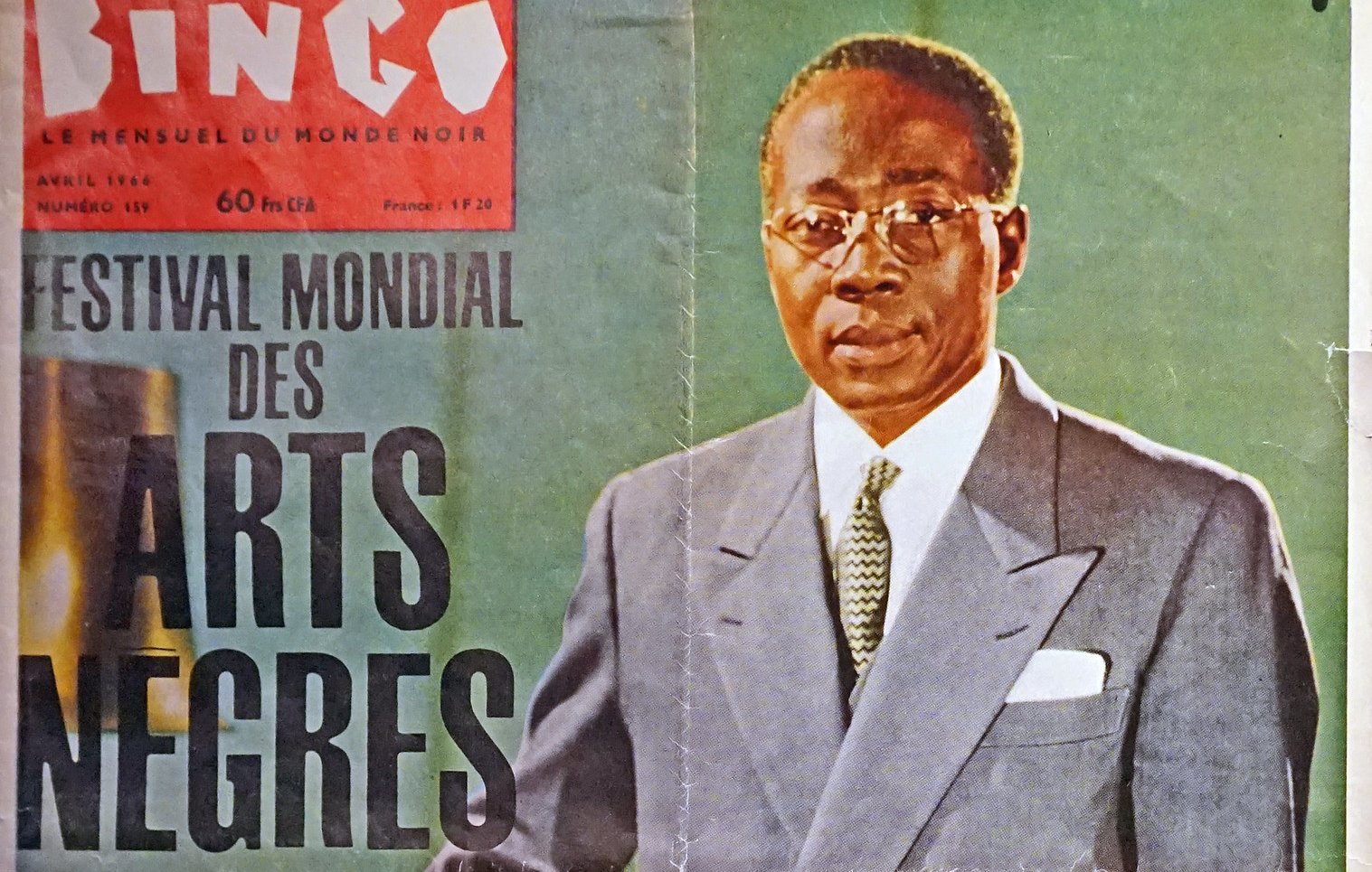 L'image représente une couverture de magazine intitulée "Bingo", datée d'avril 1966. Au premier plan, un homme debout est habillé d'un costume clair, avec une chemise blanche et une cravate sombre. Il porte des lunettes et affiche une expression sérieuse. L'arrière-plan est flou, mais semble être un environnement sobre, mettant en valeur le sujet. En haut de la couverture, on peut lire "FESTIVAL MONDIAL DES ARTS NÈGRES", ce qui évoque un événement culturel important. Les couleurs du magazine sont douces, ajoutant une touche d'élégance à l'ensemble.