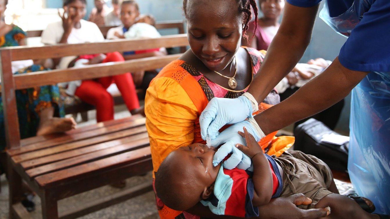 Dans cette image, nous voyons une scène dans une salle d'attente où plusieurs personnes sont assises sur des bancs en bois. Au premier plan, une mère, vêtue d'une robe orange, tient son bébé. Elle semble attentive et douce. Un professionnel de la santé, portant des gants bleus, est en train de donner un soin ou un médicament au bébé. Le bébé, habillé d'un t-shirt bleu et d'un short, a une expression calme. À l'arrière-plan, d'autres femmes et enfants attendent, créant une atmosphère de communauté et de soutien. Les murs sont peints en bleu, ajoutant une ambiance sereine à la scène.