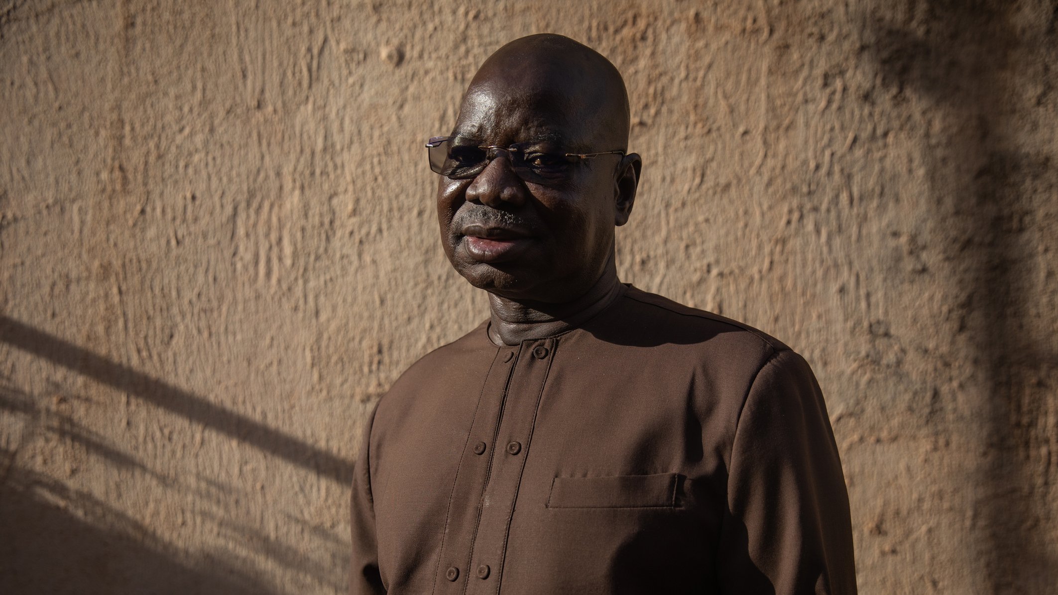 L'image présente un homme d'âge mûr, avec un teint foncé et des lunettes. Il porte une tenue sombre, légèrement scintillante, adaptée à un environnement formel ou culturel. Son expression est sereine et réfléchie. Le fond est constitué d'un mur en terre ou en adobe, éclairé par une lumière naturelle douce, créant une ambiance chaleureuse. Des ombres longues suggèrent qu'il est en fin de journée, ajoutant à la profondeur de l'image.