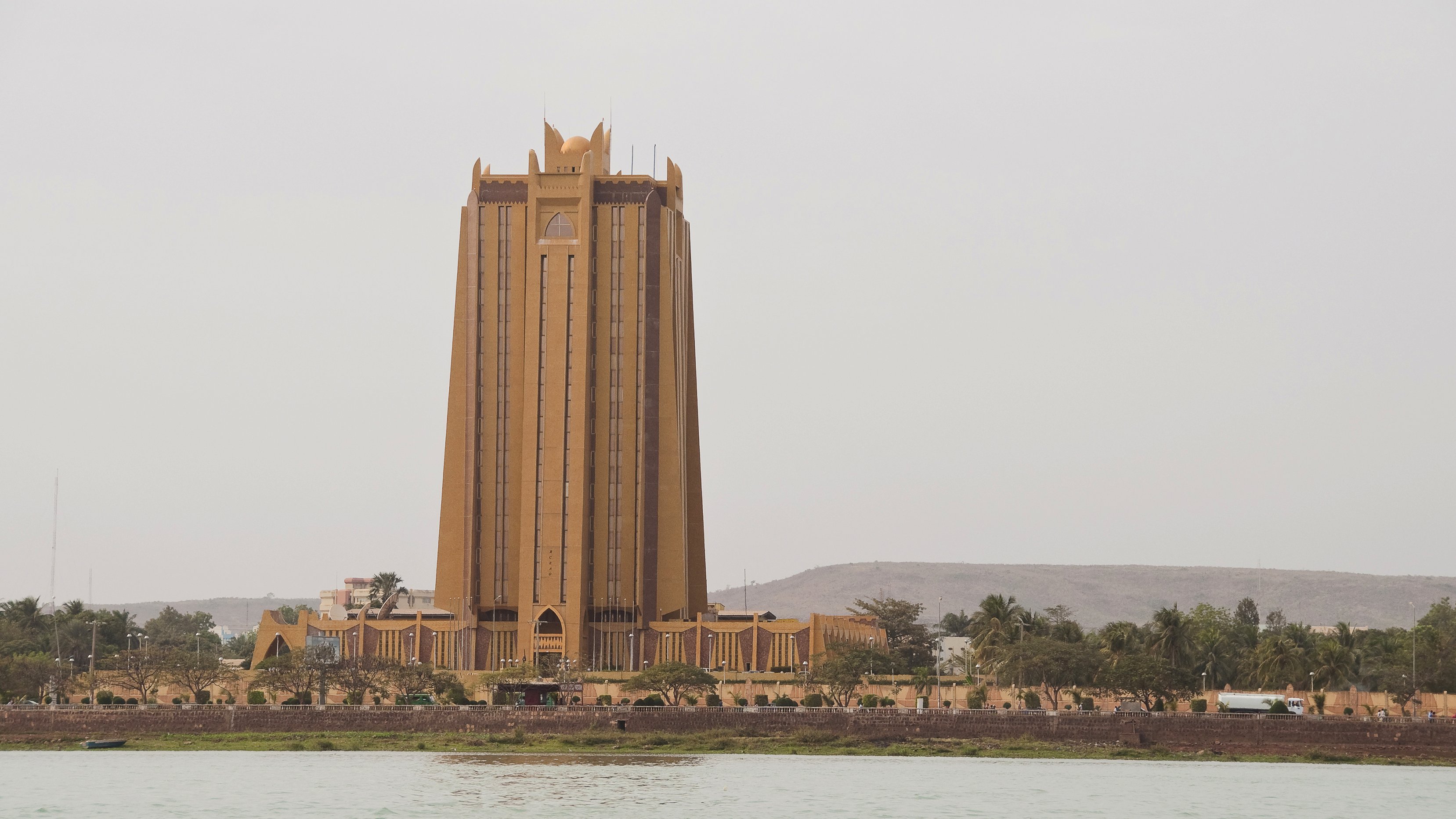 L'image montre un bâtiment imposant qui semble être une tour, située près de l'eau. Ce bâtiment, d'une teinte dorée, a une architecture moderne et élancée. Sa structure se soulève vers le ciel, avec des lignes verticales marquées et des détails décoratifs au sommet, donnant une impression de grandeur. En arrière-plan, on aperçoit quelques collines et des arbres, tandis qu'au premier plan, on voit la surface calme de l'eau. Le ciel est légèrement nuageux, créant une ambiance tranquille et légèrement brumeuse autour de cette scène urbaine.